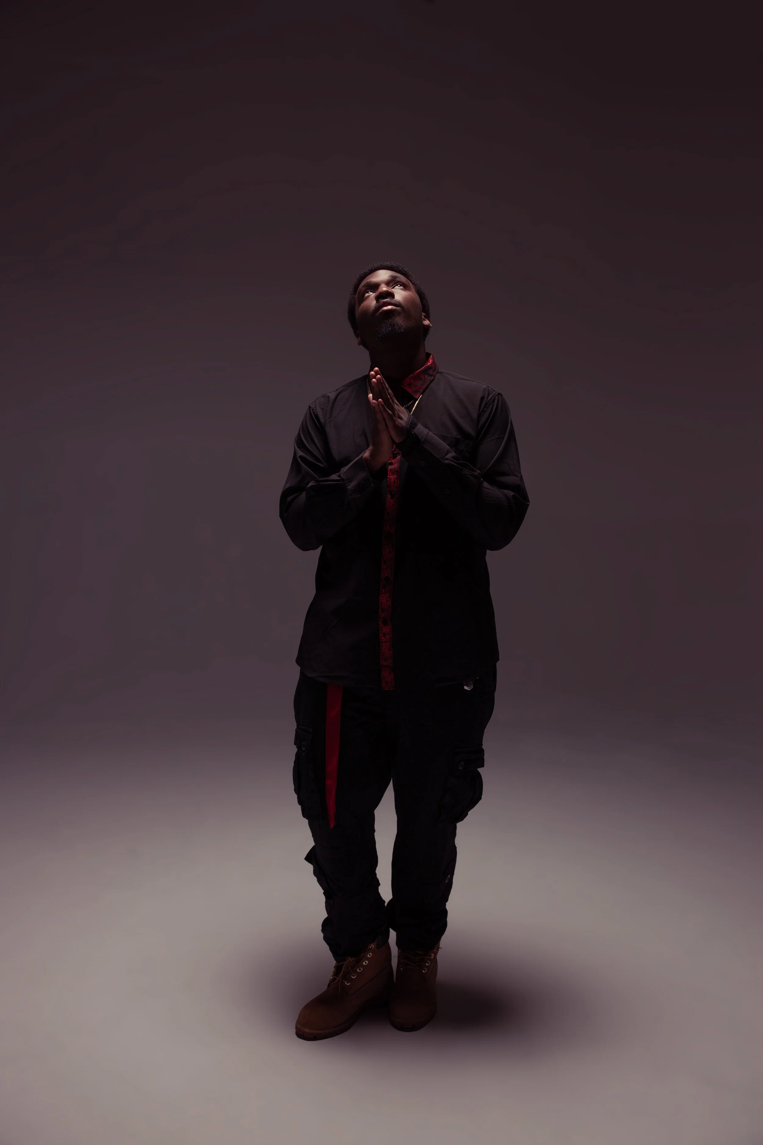 A man dressed in dark clothing, including a jacket, cargo pants, and boots, standing with hands clasped in prayer and looking upward against a plain, dark background.