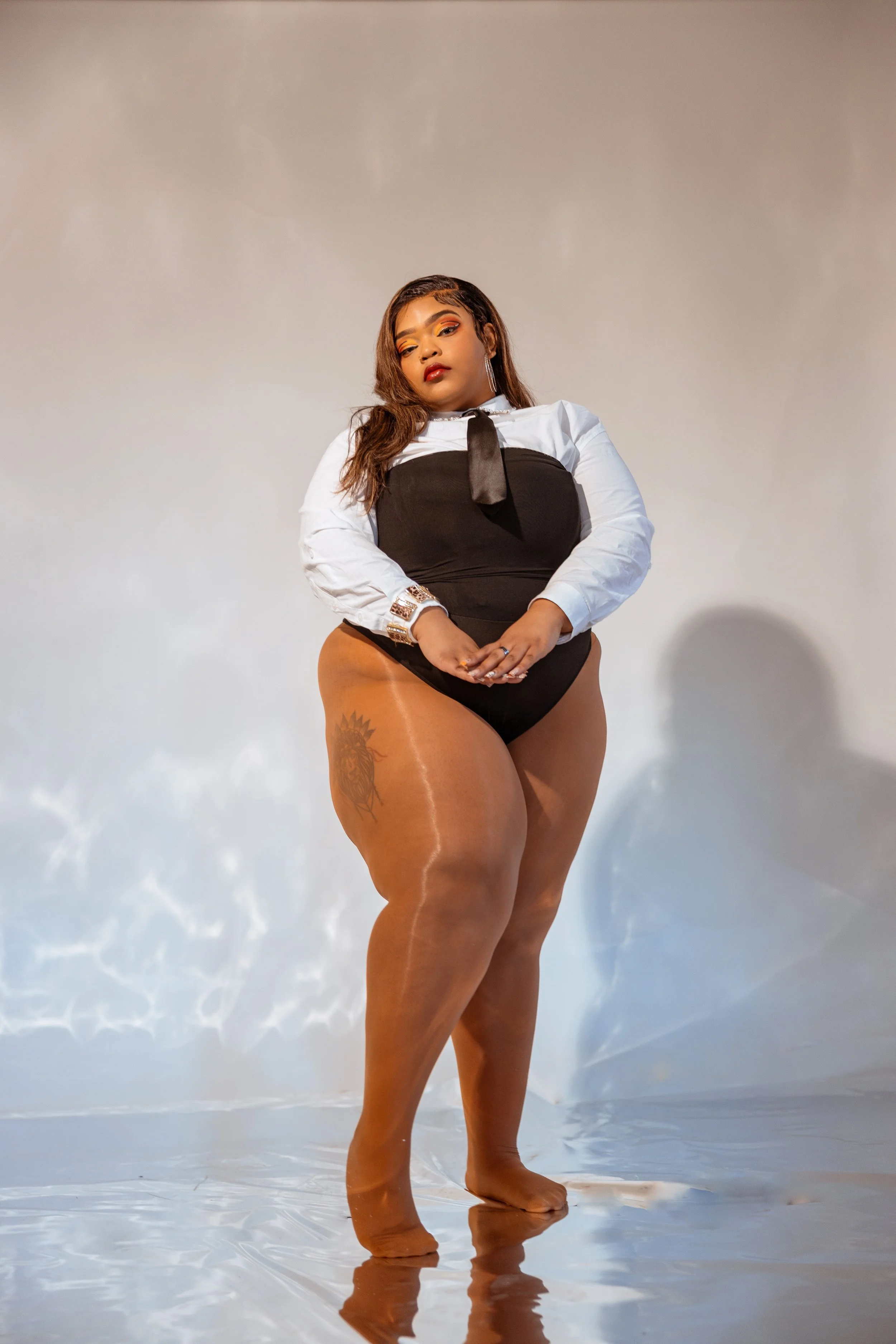 A woman standing on a reflective surface against a plain background, wearing a black and white outfit, with long brown hair and tattoos on her thigh.