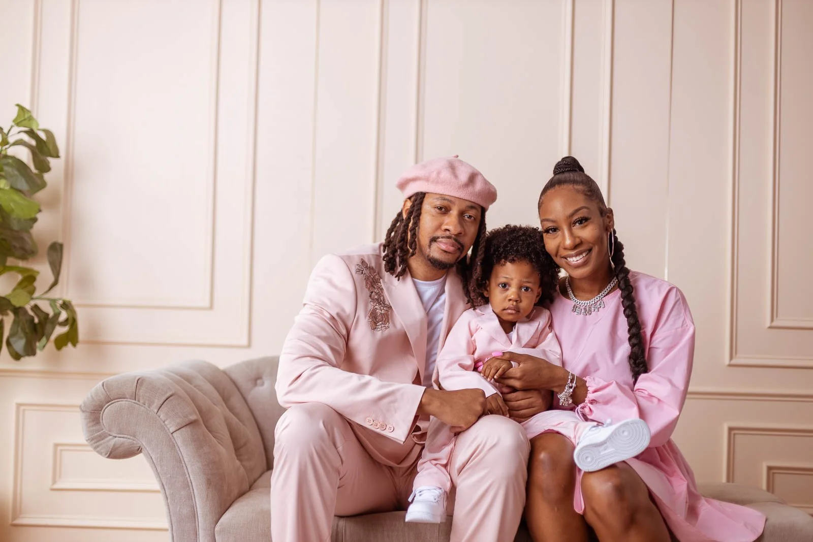 A family of three sitting on a beige sofa in a stylish, neutral-colored room. The father has dreadlocks, a pink beret, and a pink jacket. The mother has braided hair, a large smile, and wears a pink dress. Their young daughter, with curly hair, sits 