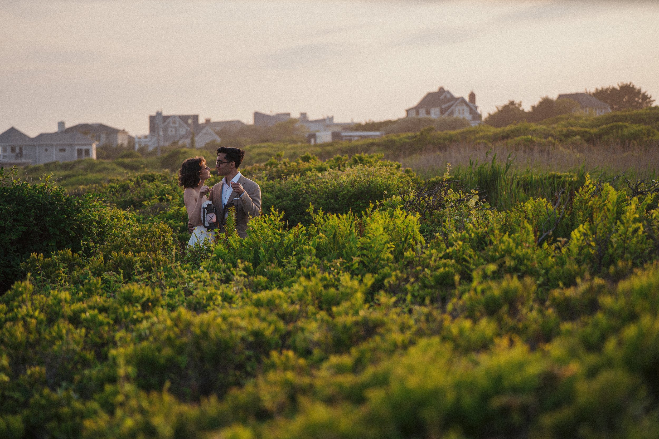 A young couple stands close together in a field of green bushes, with houses on a hill in the background, during sunset.