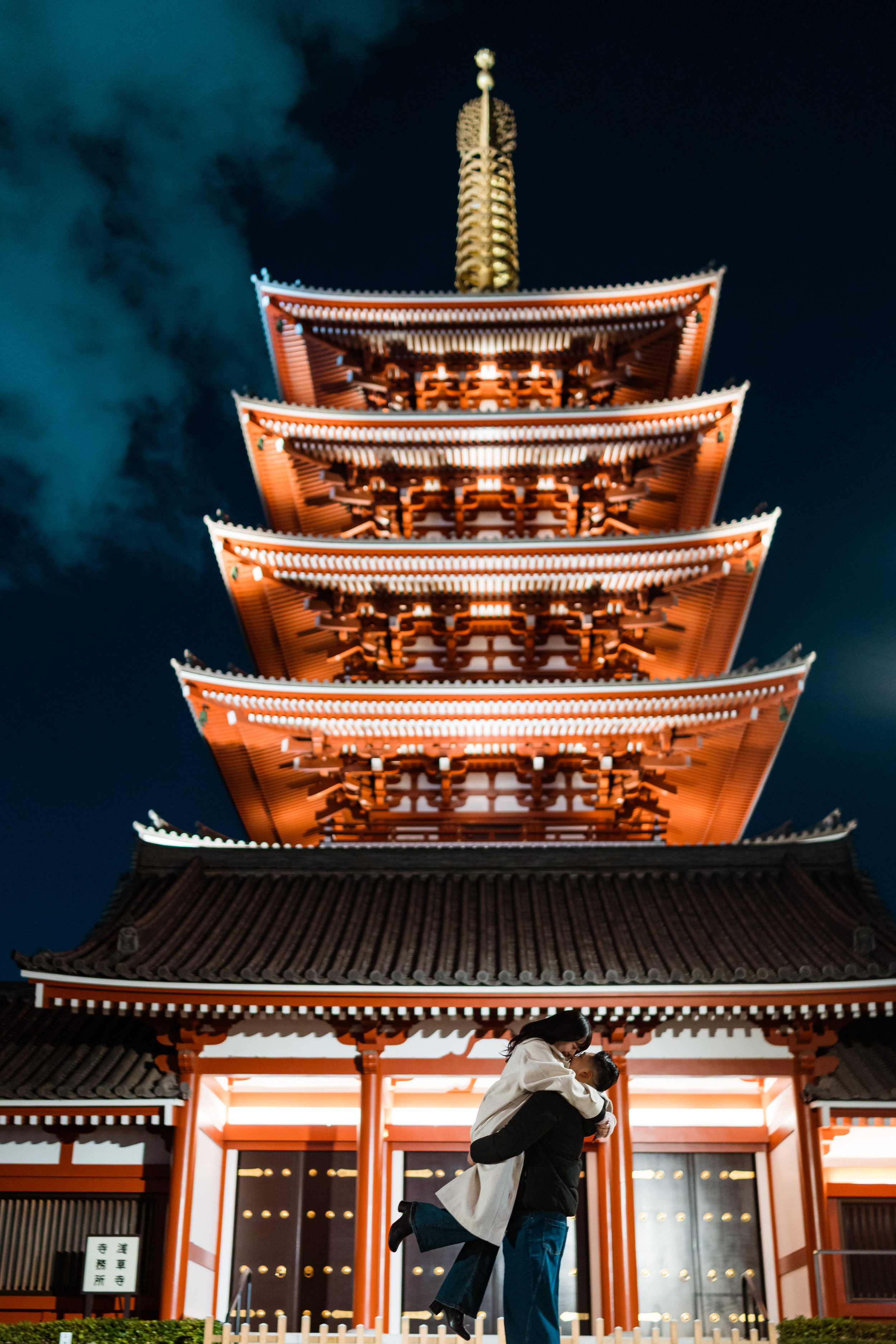 A couple embraces in front of a illuminated traditional Japanese pagoda at night.