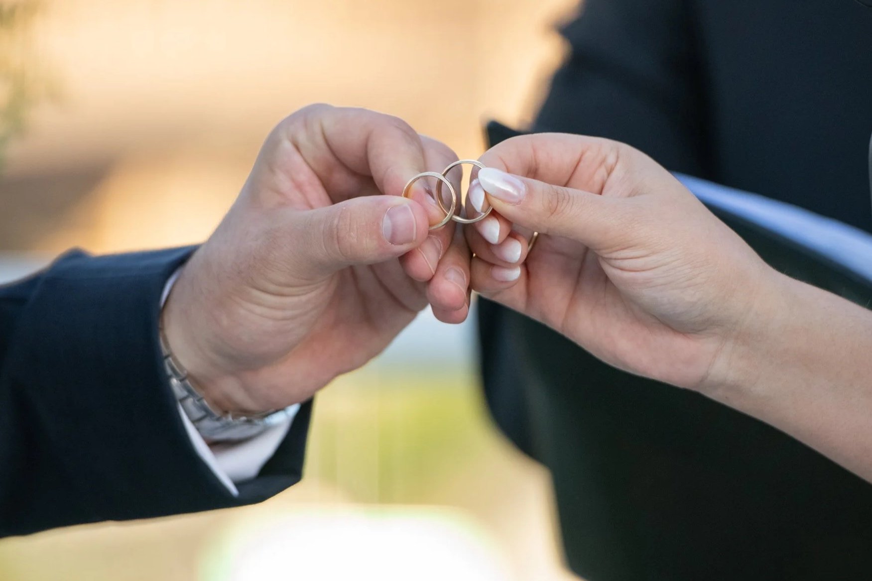A person in a black suit is handing a gold wedding band to a woman in a wheelchair during an outdoor ceremony.