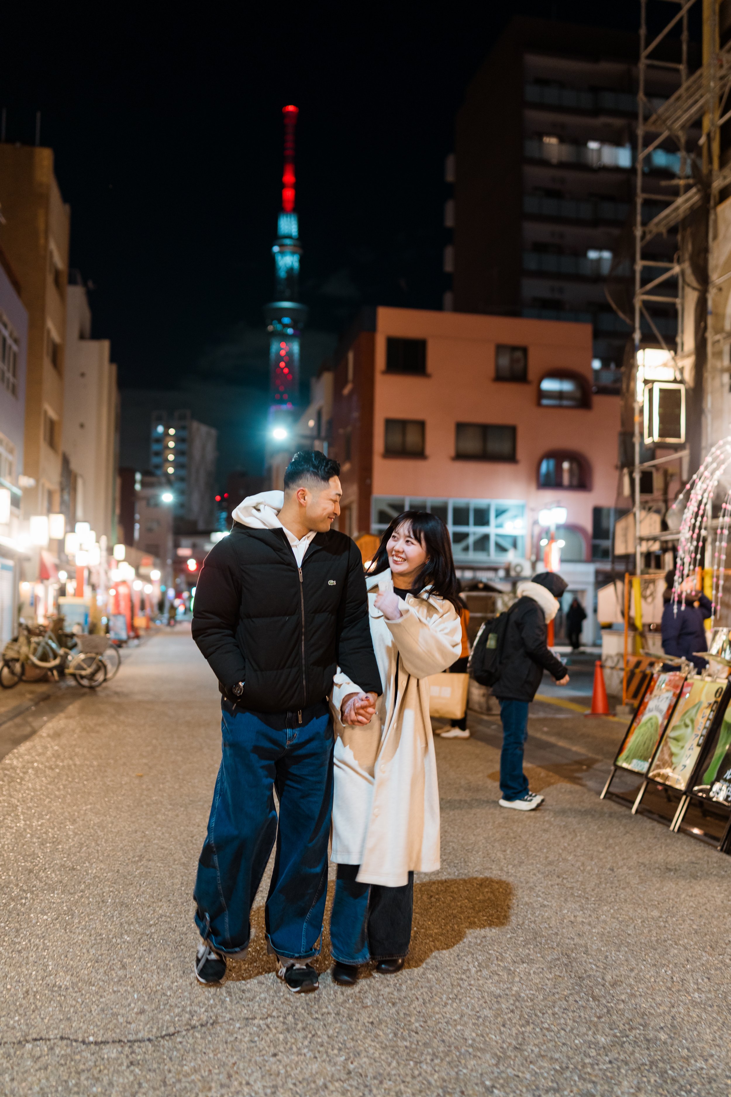 A young couple holding hands and smiling on a city street at night, with street vendors and the Tokyo Skytree in the background.