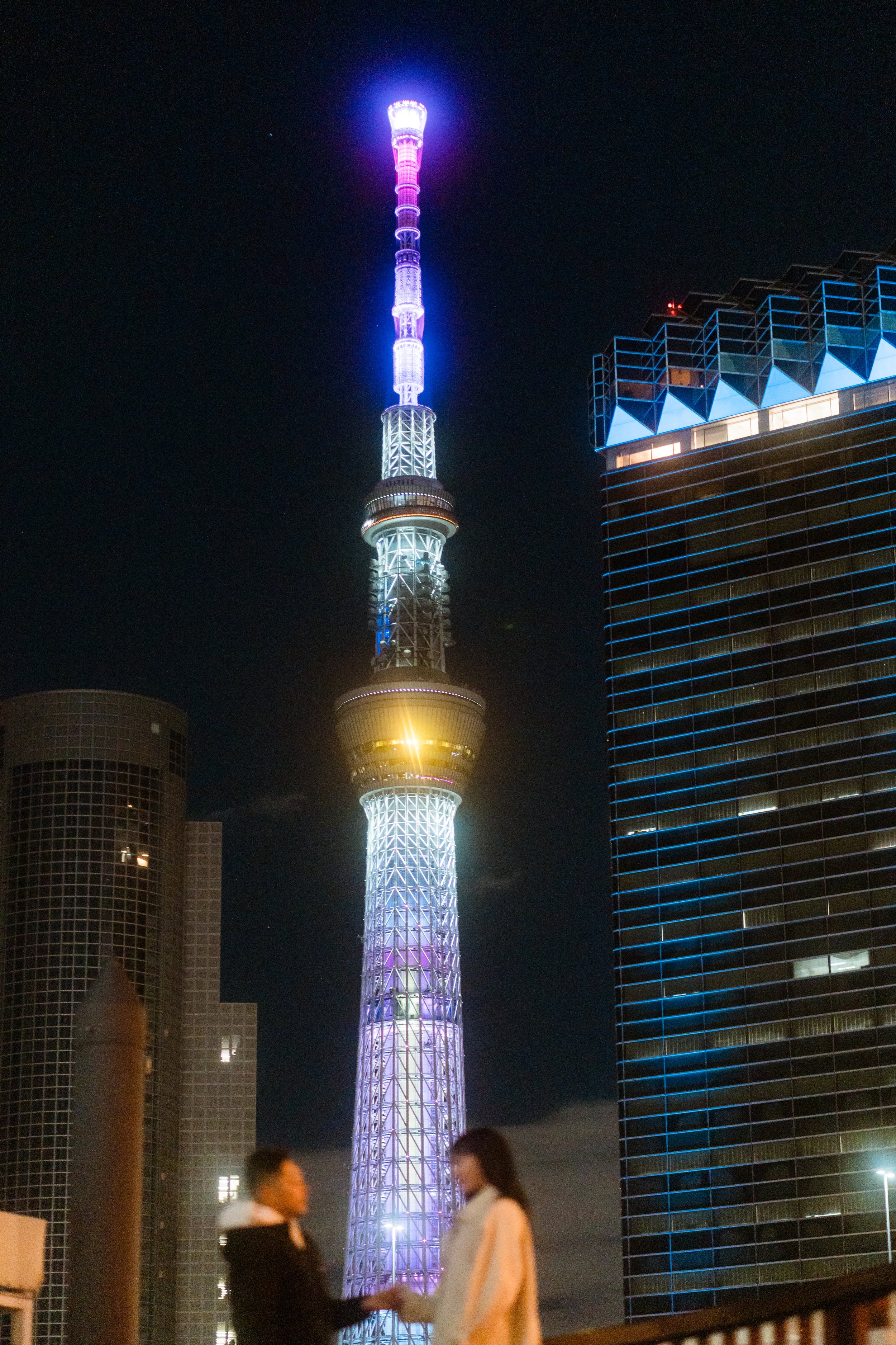Nighttime view of Tokyo Skytree with colorful lighting, two blurred people shaking hands in the foreground, and tall buildings surrounding the tower.
