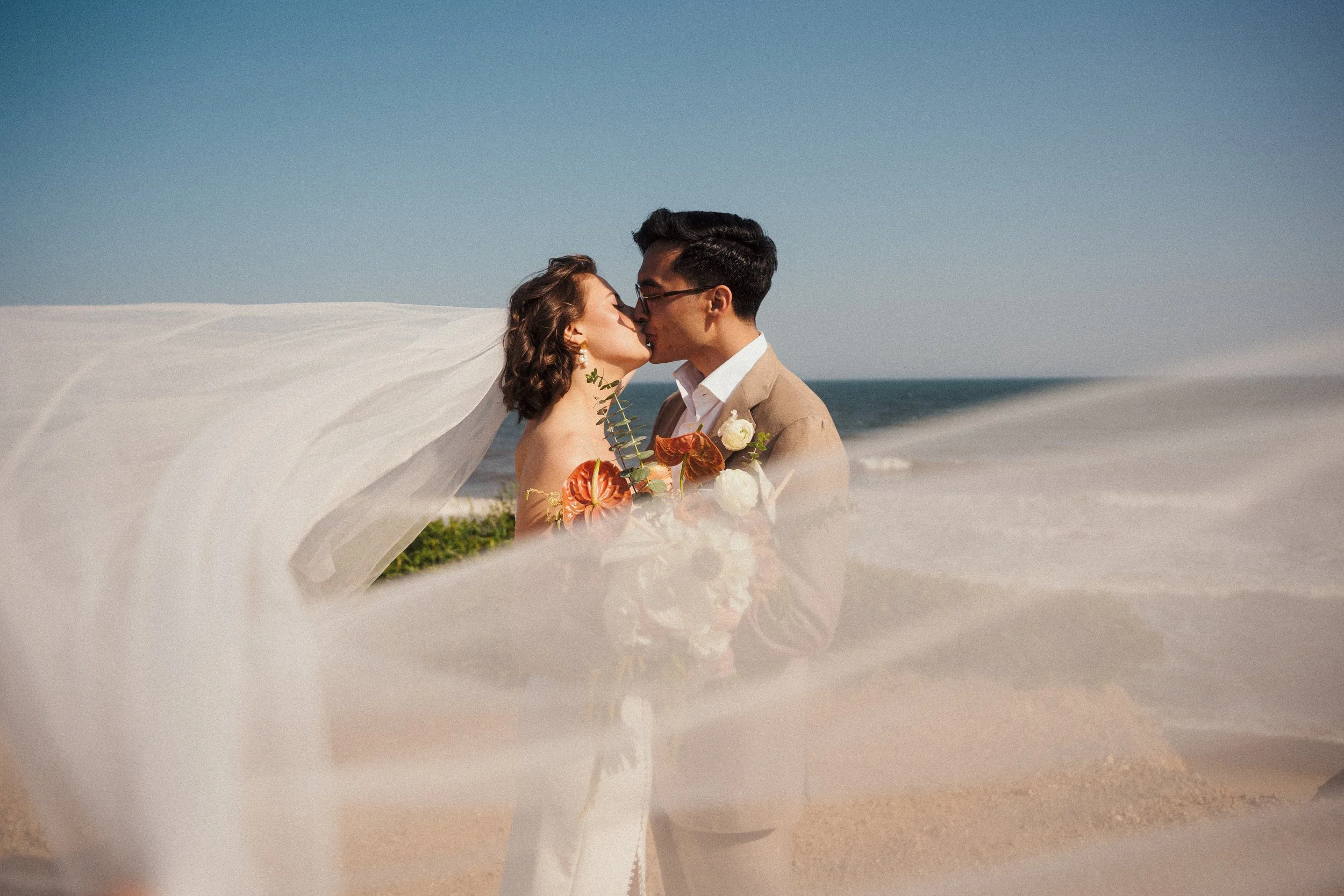 A bride and groom kissing on the beach, with the bride's veil flowing in the wind.