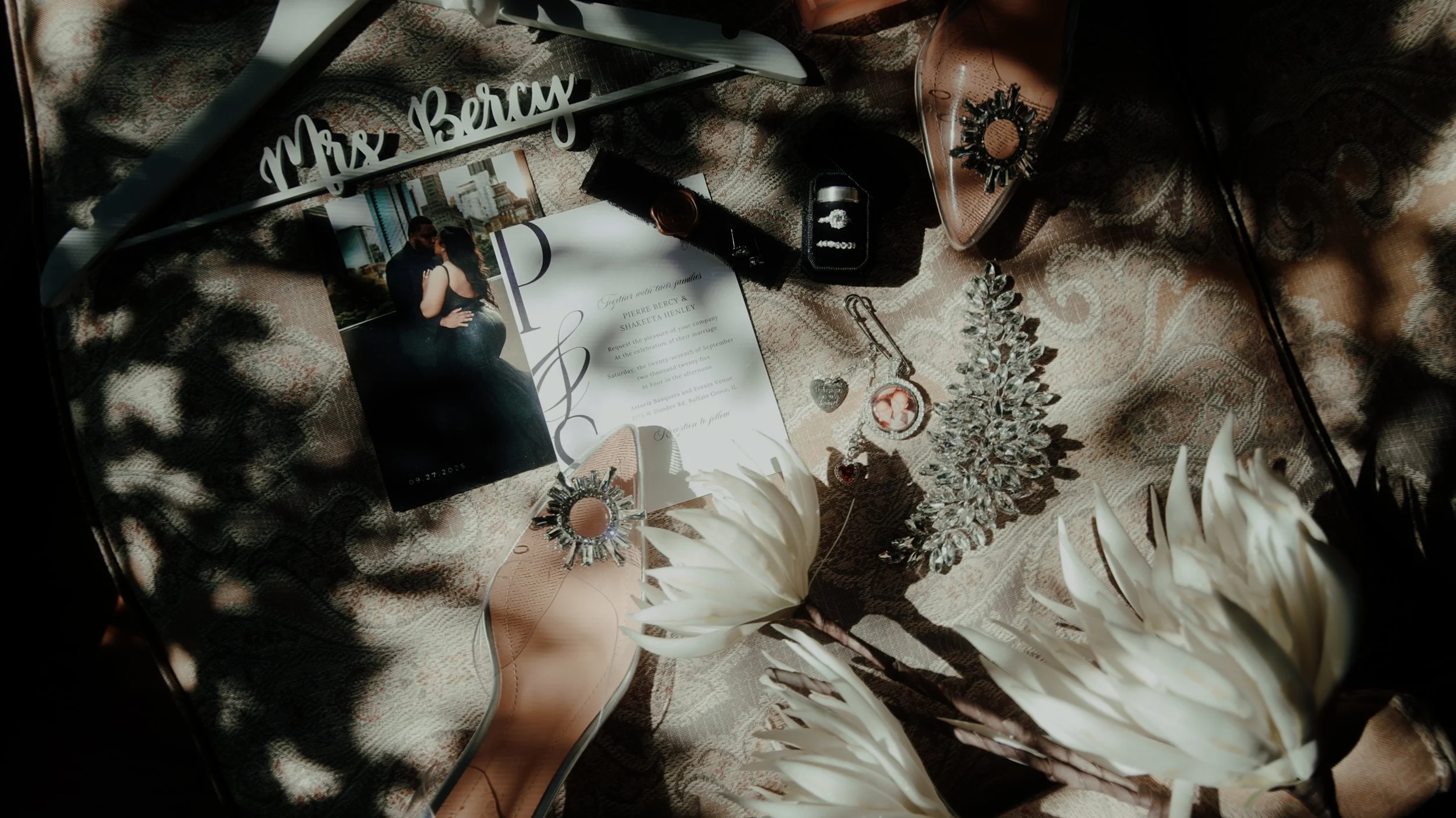 A wedding invitation, a photo of a couple, jewelry, a perfume bottle, a flower, a pair of shoes, and other decorative items on fabric.