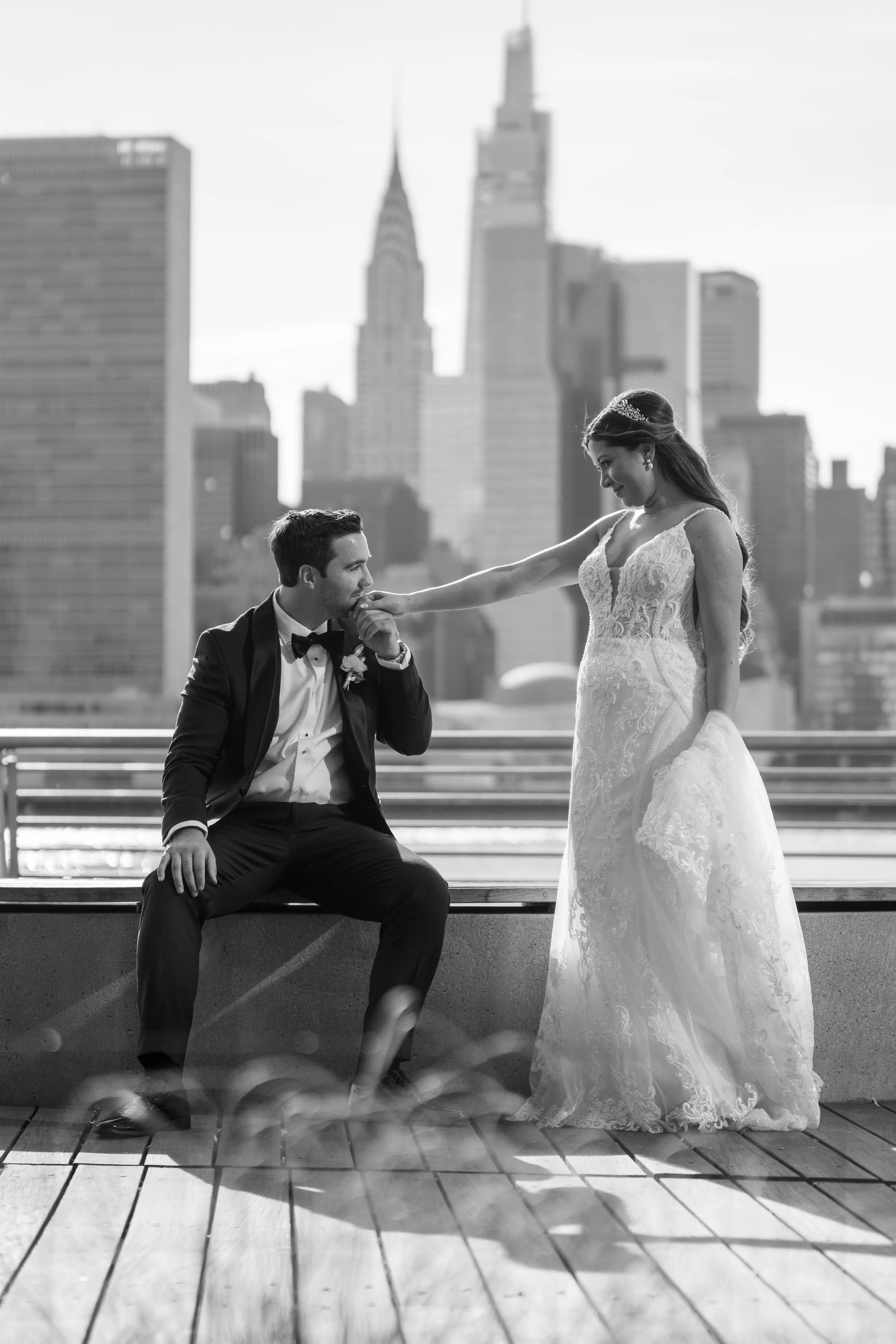 A black and white photo of a bride and groom on a rooftop. The groom, sitting on a bench, is kissing the bride's hand while she stands next to him, leaning slightly towards him. The bride is wearing a detailed lace wedding dress with a tiara, and the