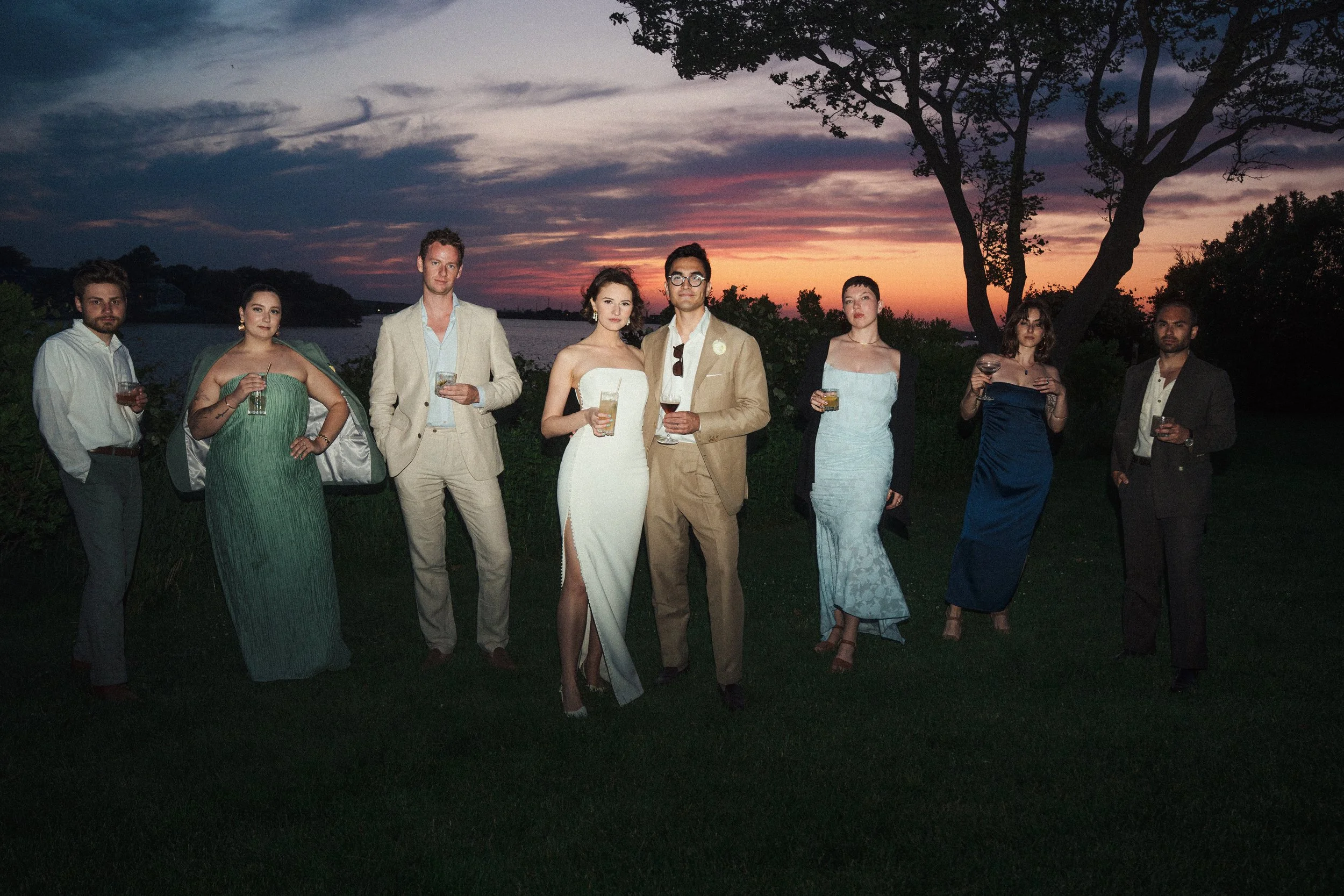 A group of eight people dressed in formal attire standing outdoors during sunset, holding drinks.
