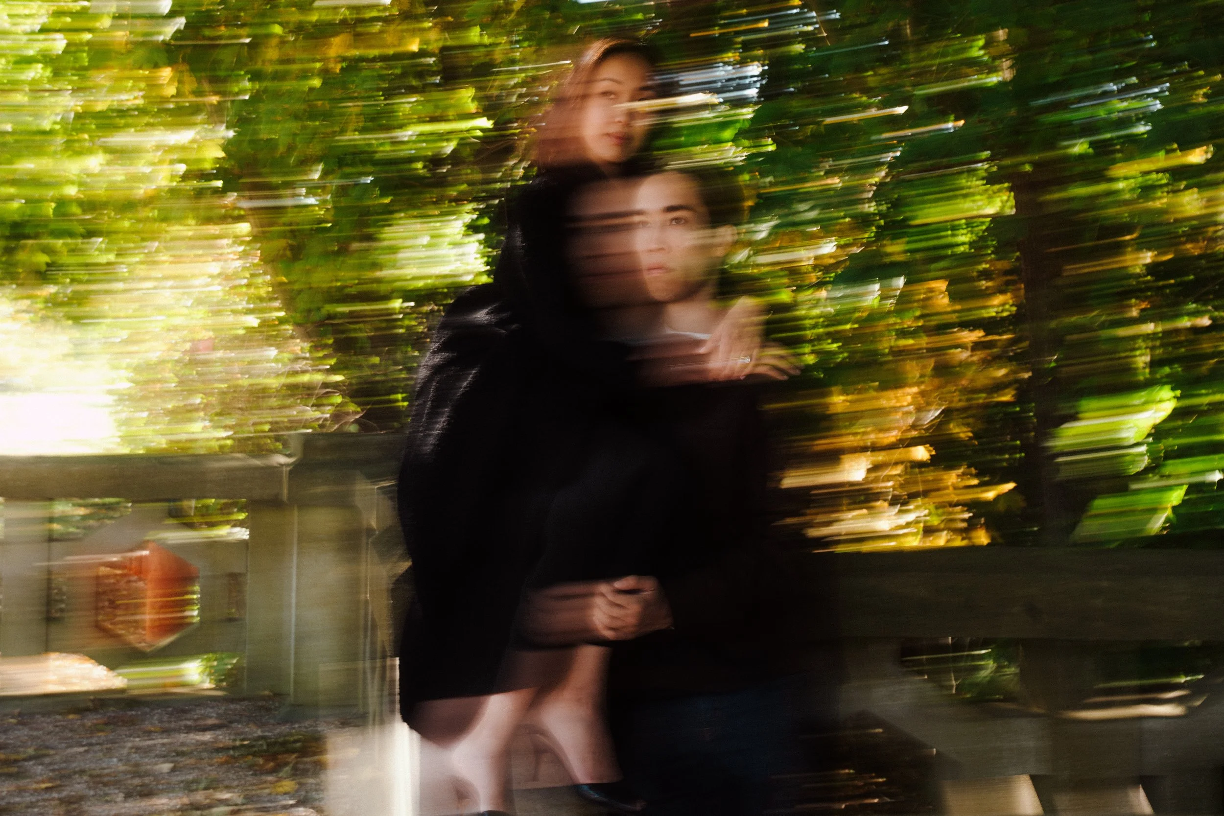 Blurred image of two women, one holding a toddler, standing outdoors on a wooden bridge with green foliage background.