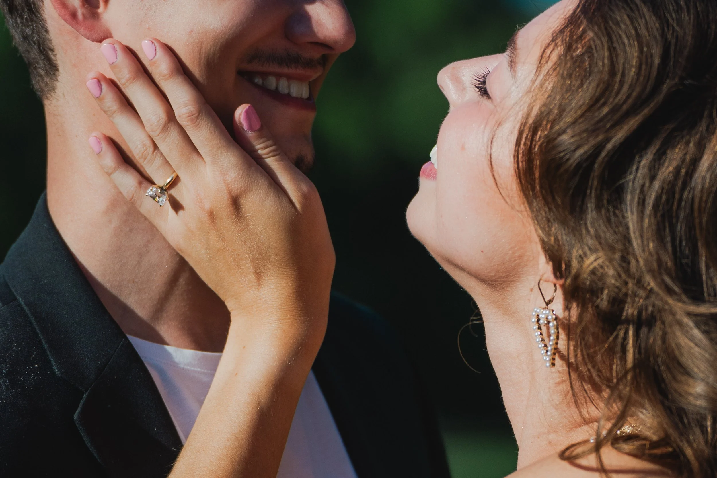 A close-up of a happy couple about to kiss, with the woman touching the man's face. The woman has curly hair and is wearing earrings, and the man is wearing a suit jacket. The woman has a ring on her finger.