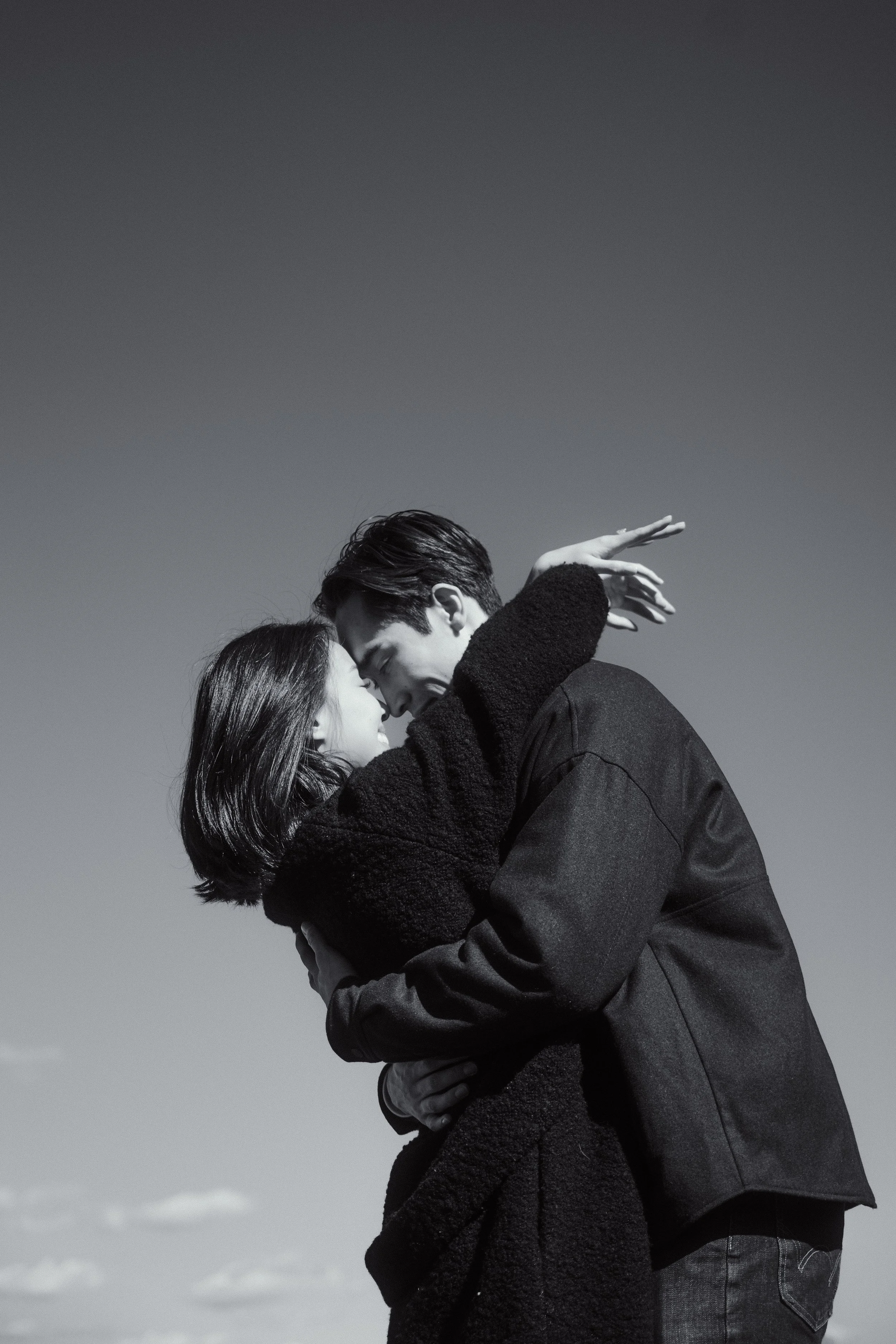 A black-and-white photo of a couple embracing outdoors against a clear sky. They are close, with faces touching, expressing affection.