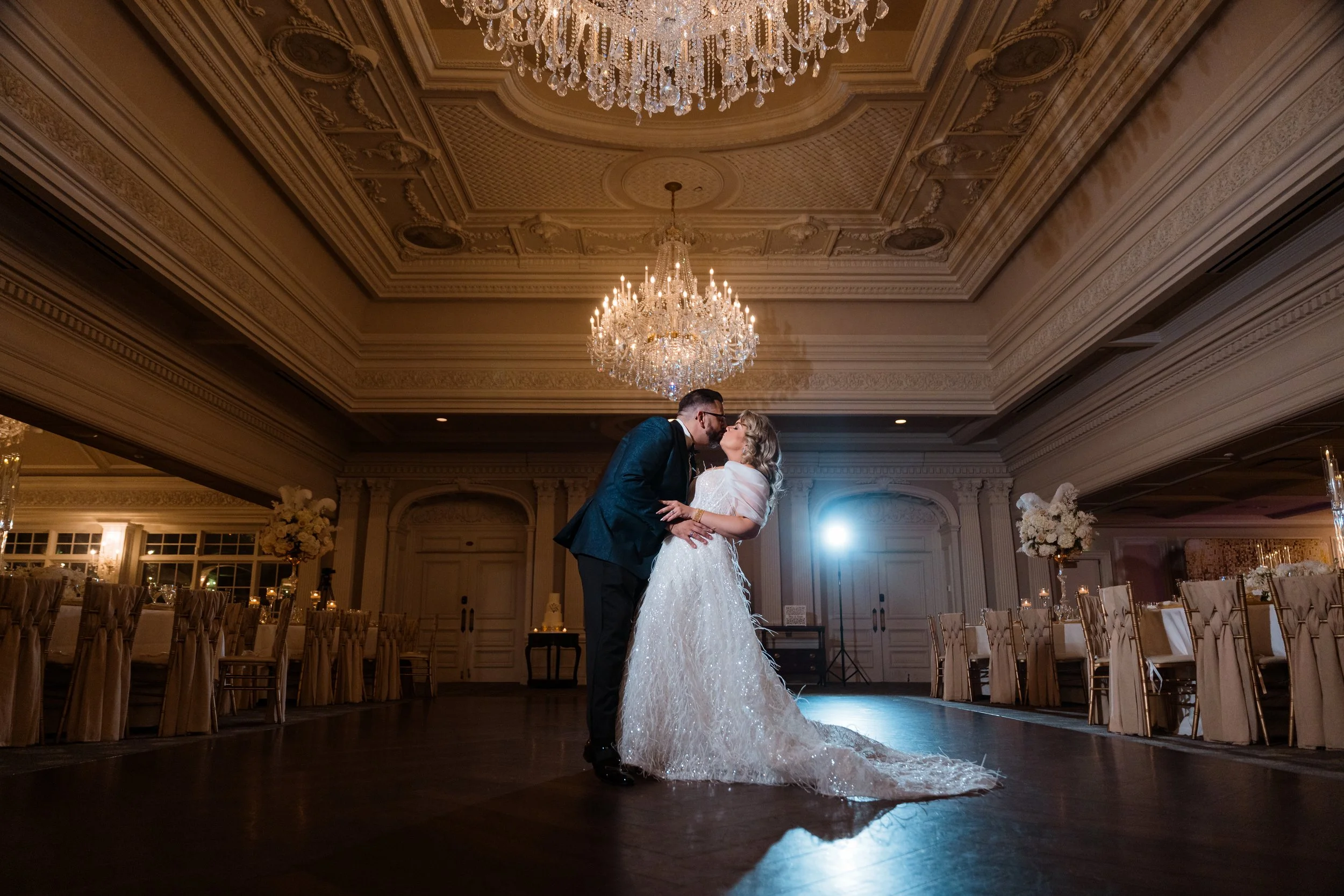 A bride and groom share a dance in a grand decorated ballroom with chandeliers and floral arrangements.
