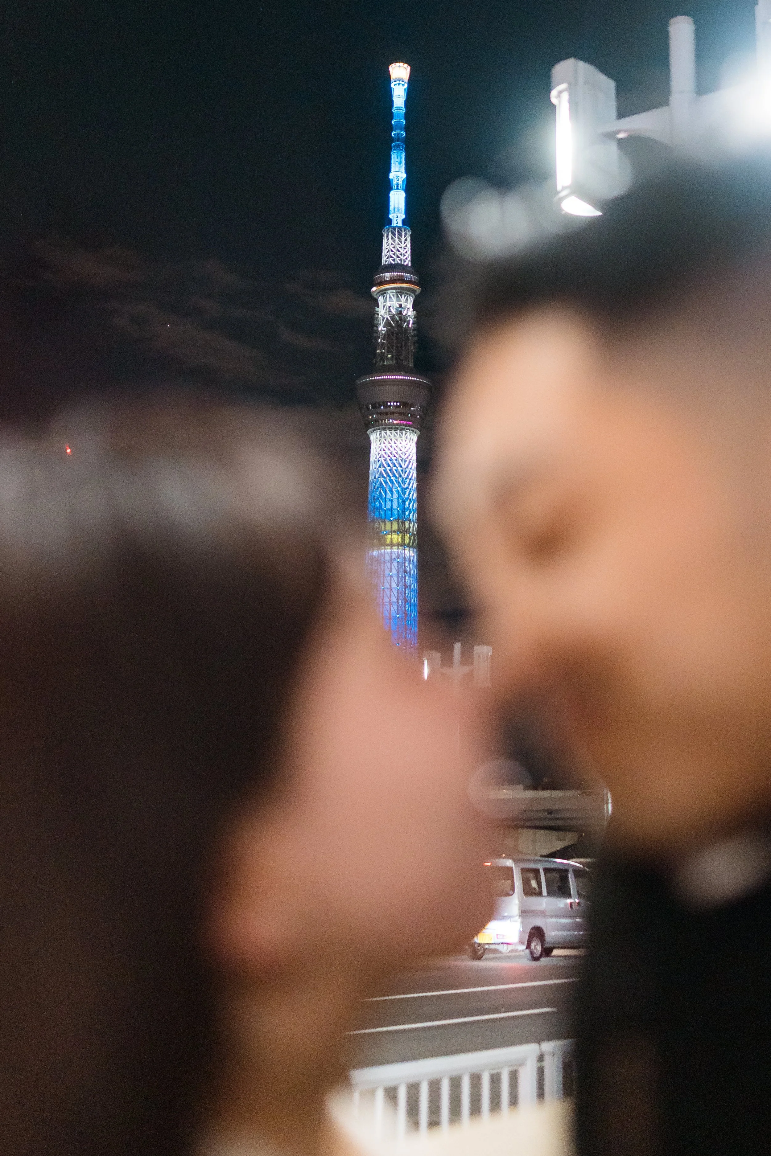 Night view of Tokyo Skytree illuminated with blue, white, and red lights, seen through the blurred outline of two people's faces.