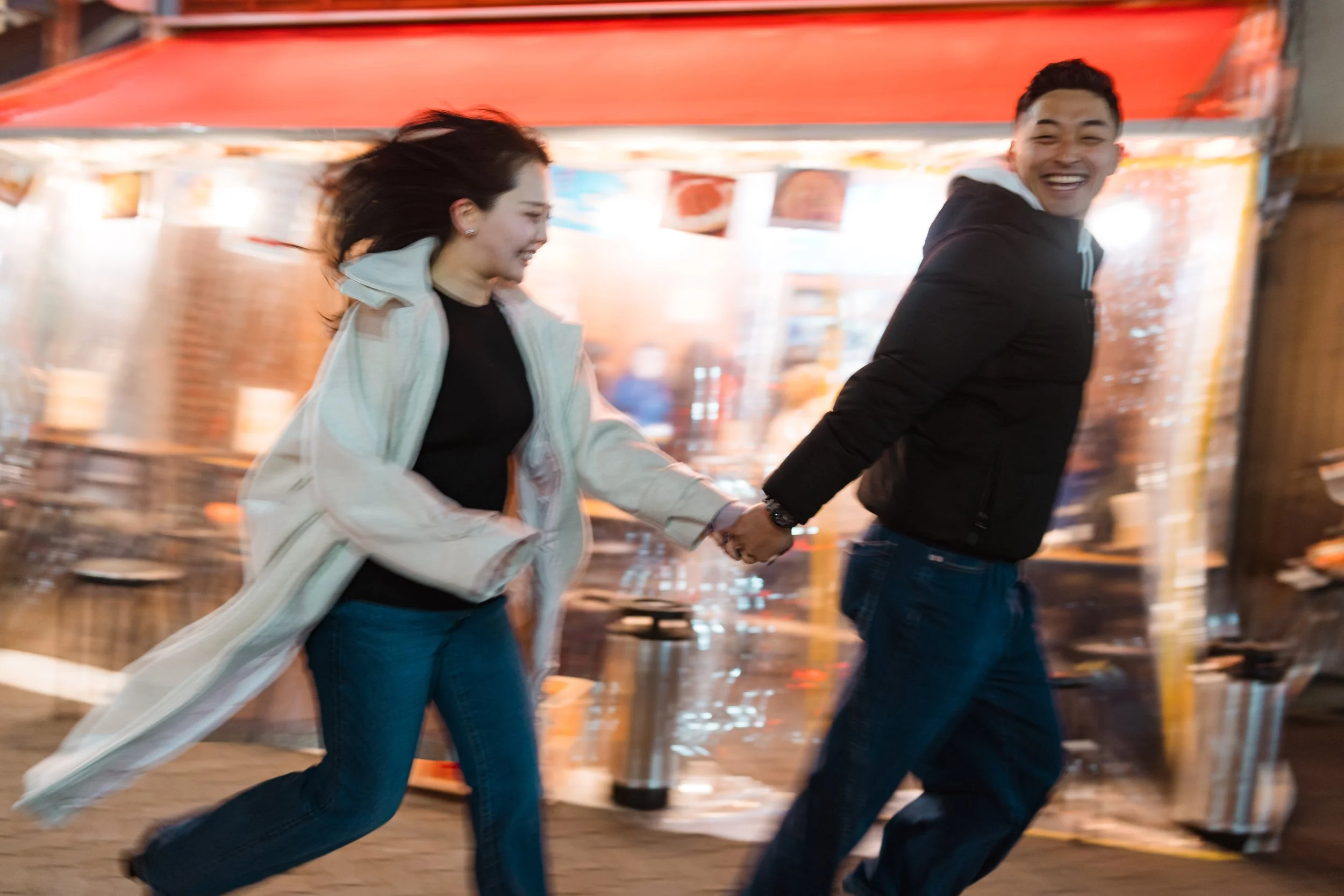 A man and woman holding hands and running through a fair or carnival at night, smiling and enjoying themselves.