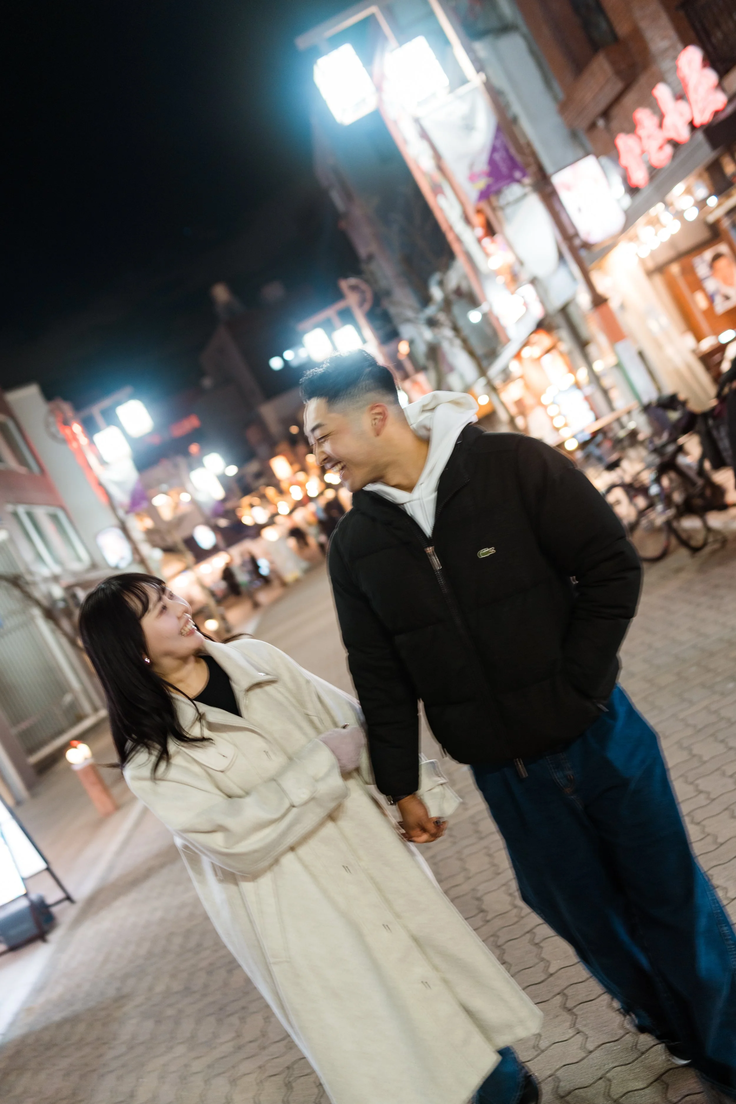 A smiling couple holding hands and walking together at night downtown, illuminated by bright city lights and neon signs.