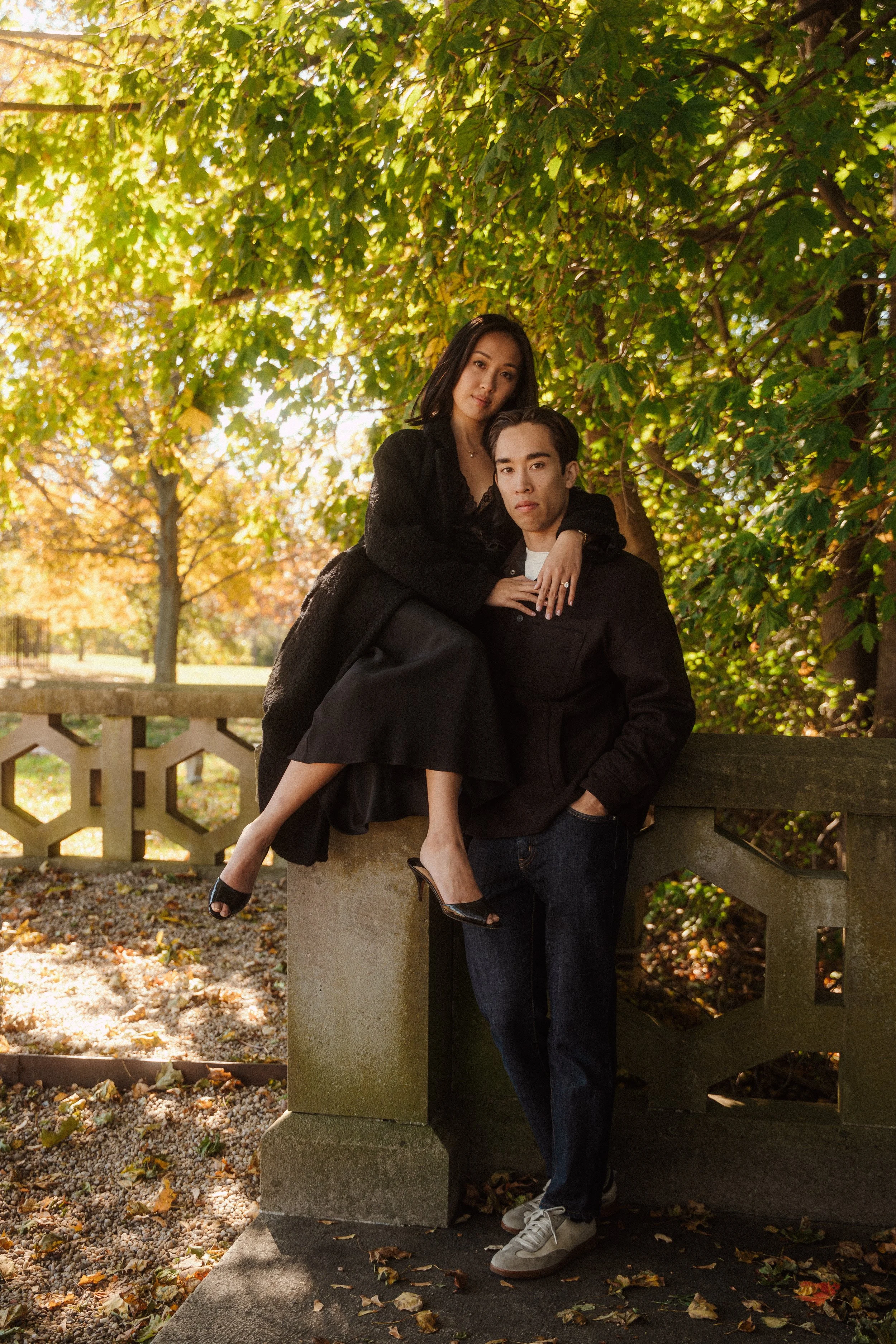 A woman sitting on a man’s lap, both dressed in black, outdoors in a park with autumn leaves and trees with green and orange foliage.