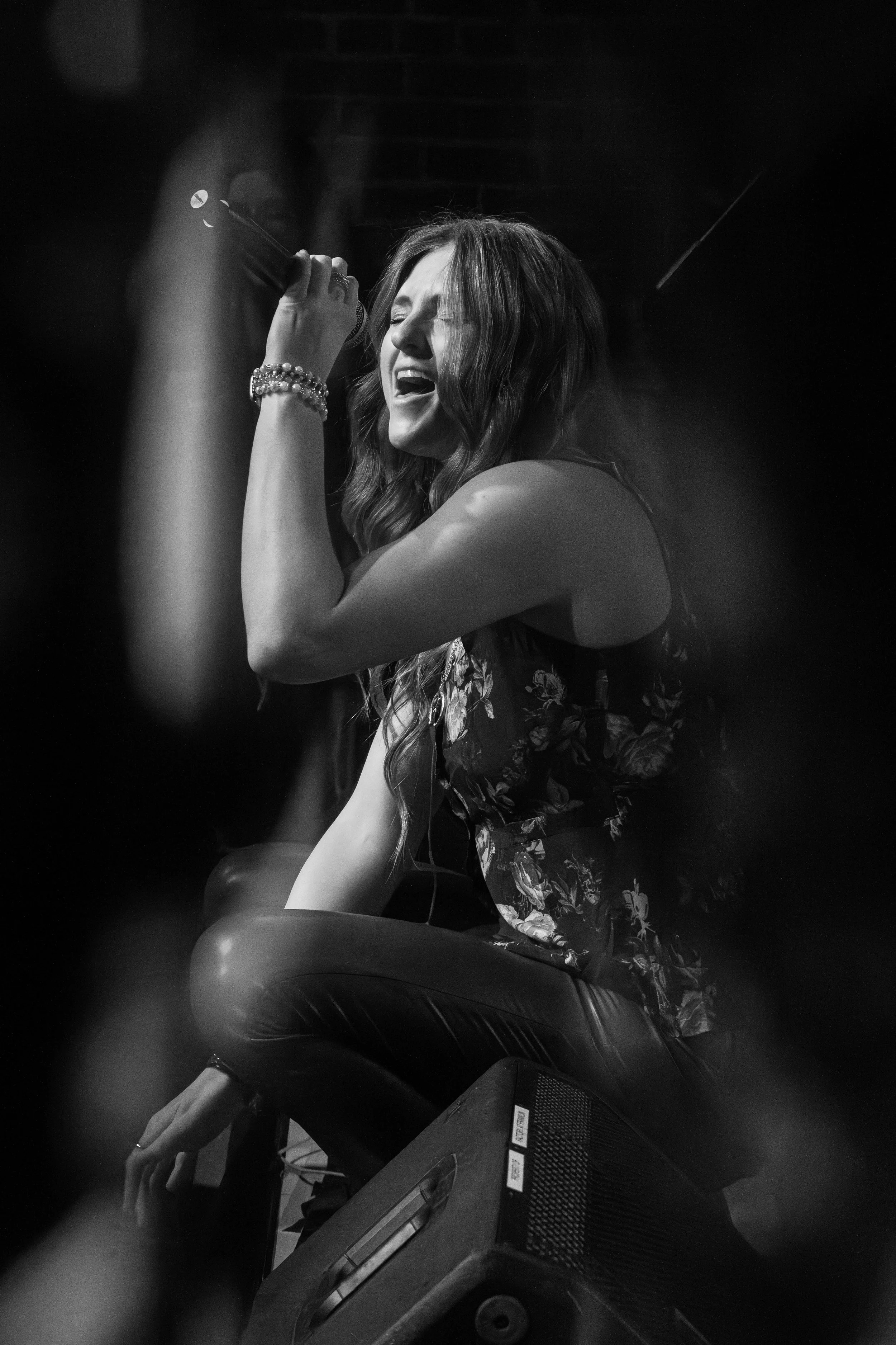 A woman with long hair is singing passionately into a microphone on stage, sitting on a speaker, wearing a sleeveless floral top and leather pants, with her eyes closed.