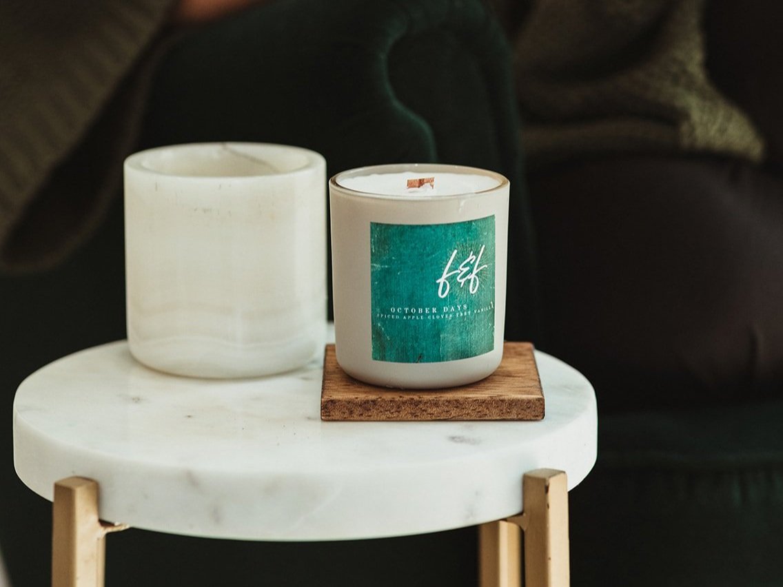 A white candle on a round marble table, next to an empty ceramic holder. The candle has a green label with text.