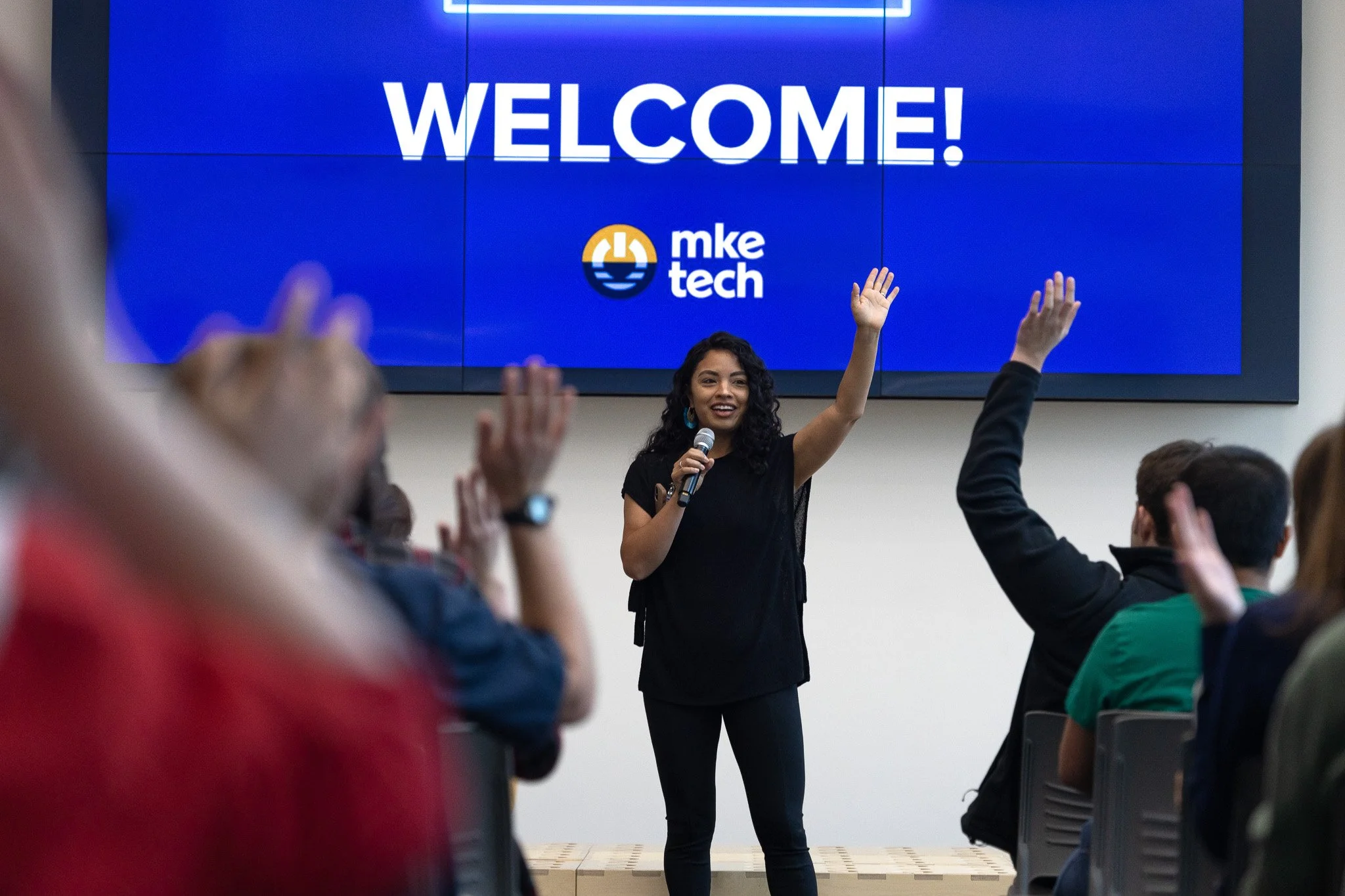 A speaker on stage greets a tech-community audience with raised hands beneath an MKE Tech welcome screen.