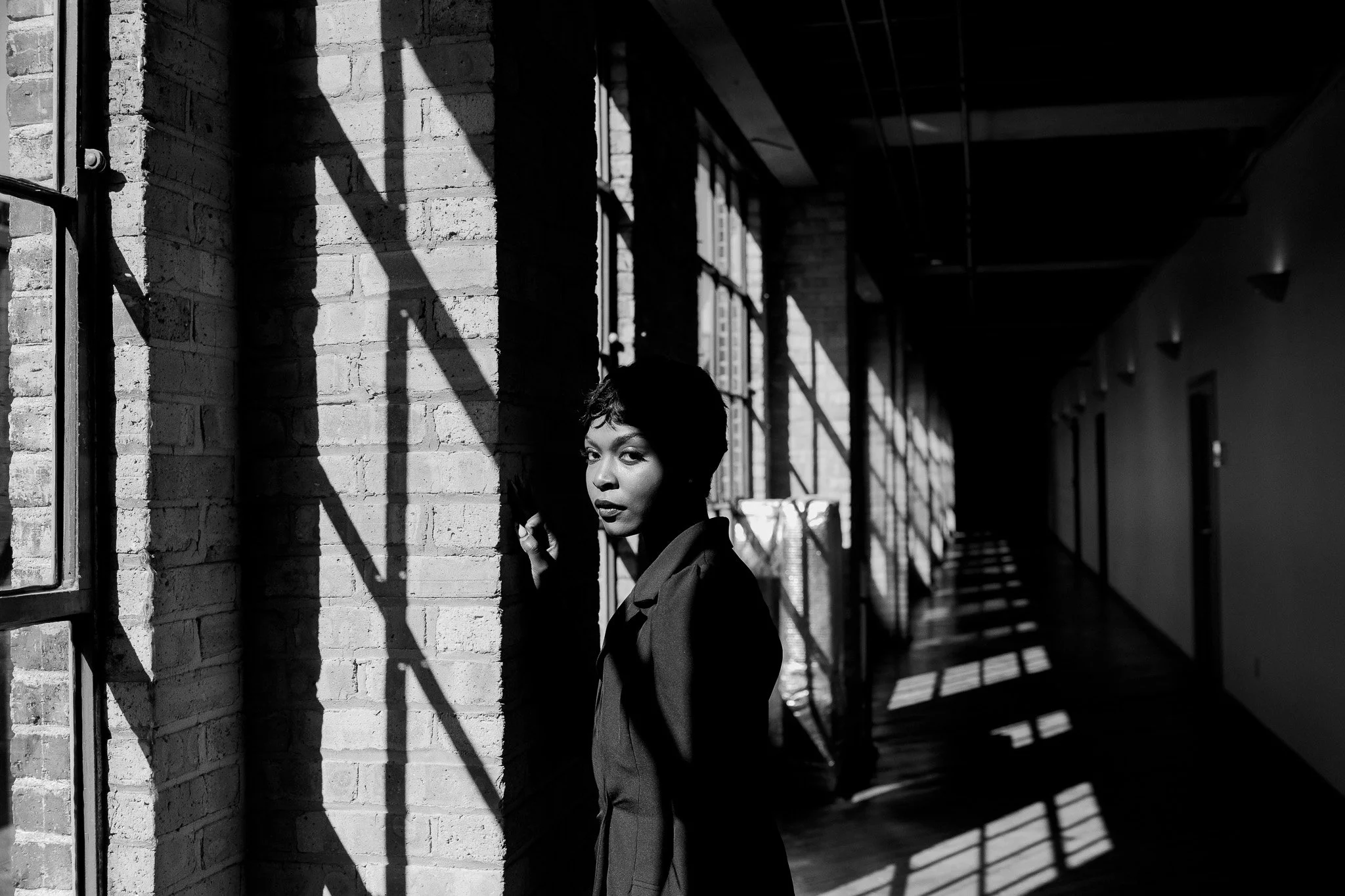 A black-and-white portrait of a woman half-turned in a sunlit brick corridor, shadows falling across the wall.