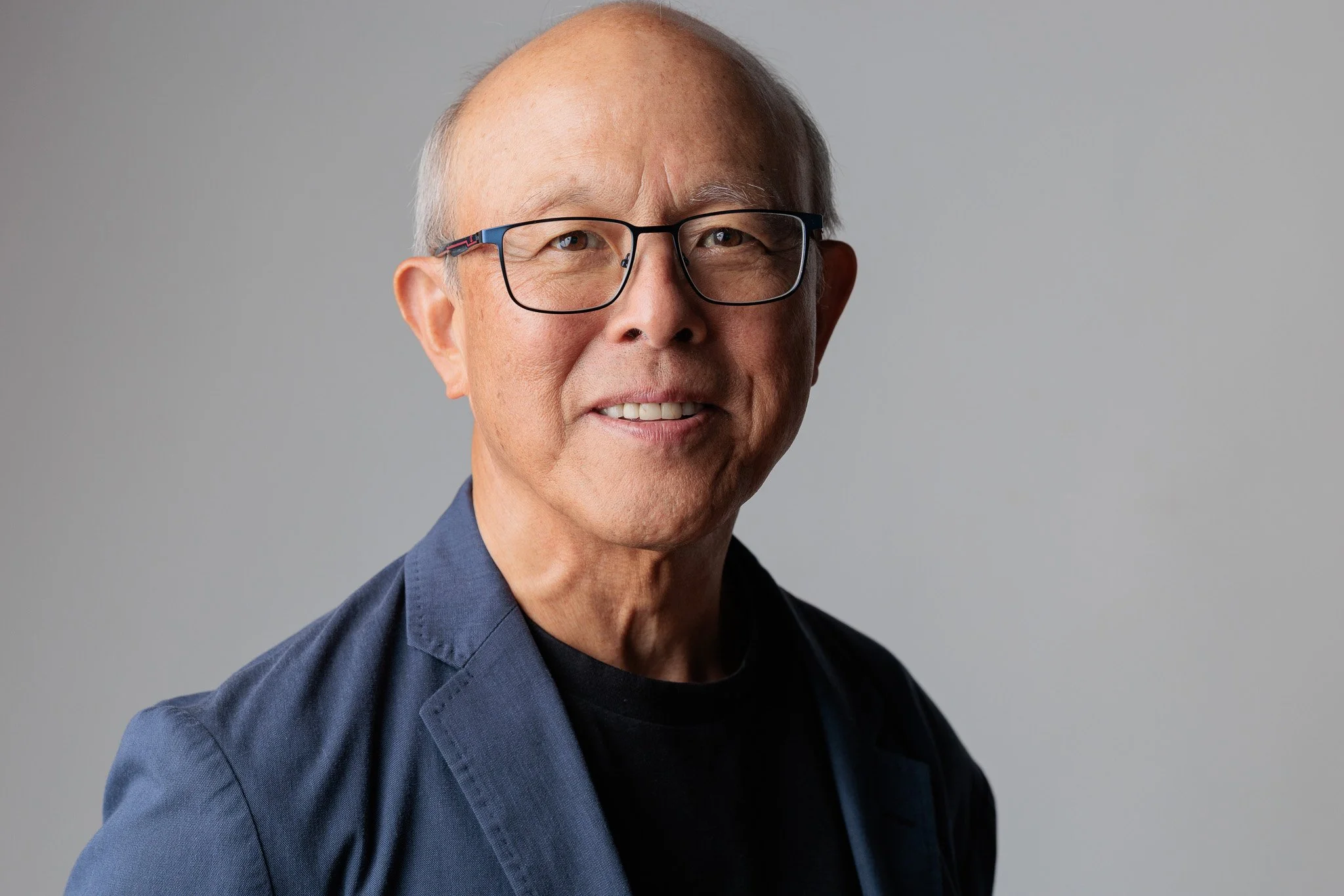A studio headshot of a smiling man in a blue blazer against a soft gray background.