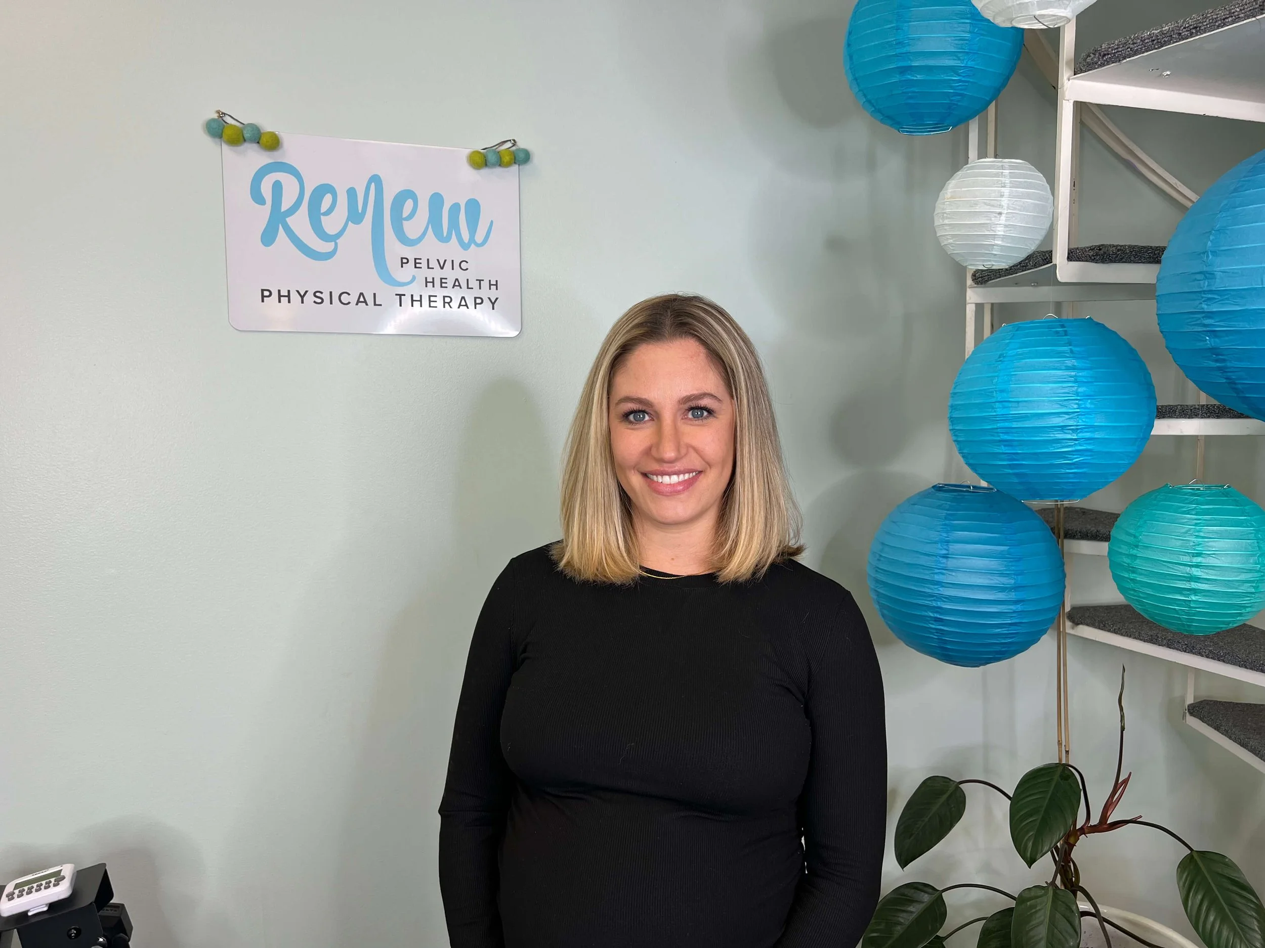 Ashley Zimmerman smiles standing in front of Renew Pelvic Health sign, blue hanging lanterns, and a plant