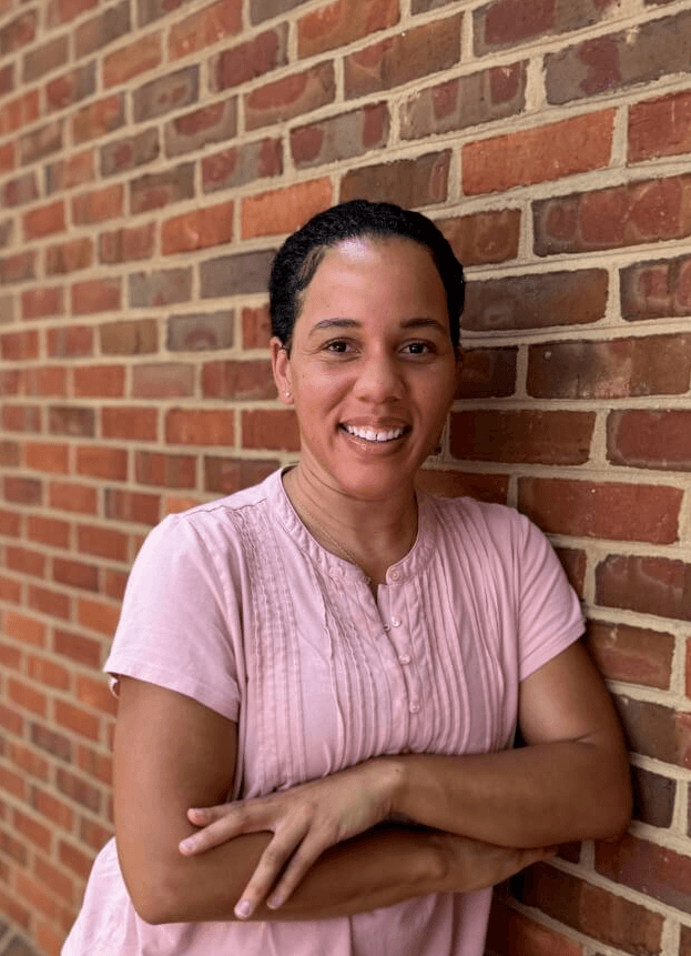 Dyane Loney pelvic floor physical therapist wears a pink shirt and smiles with her arms crossed in front of a red bring wall at Renew Pelvic Health