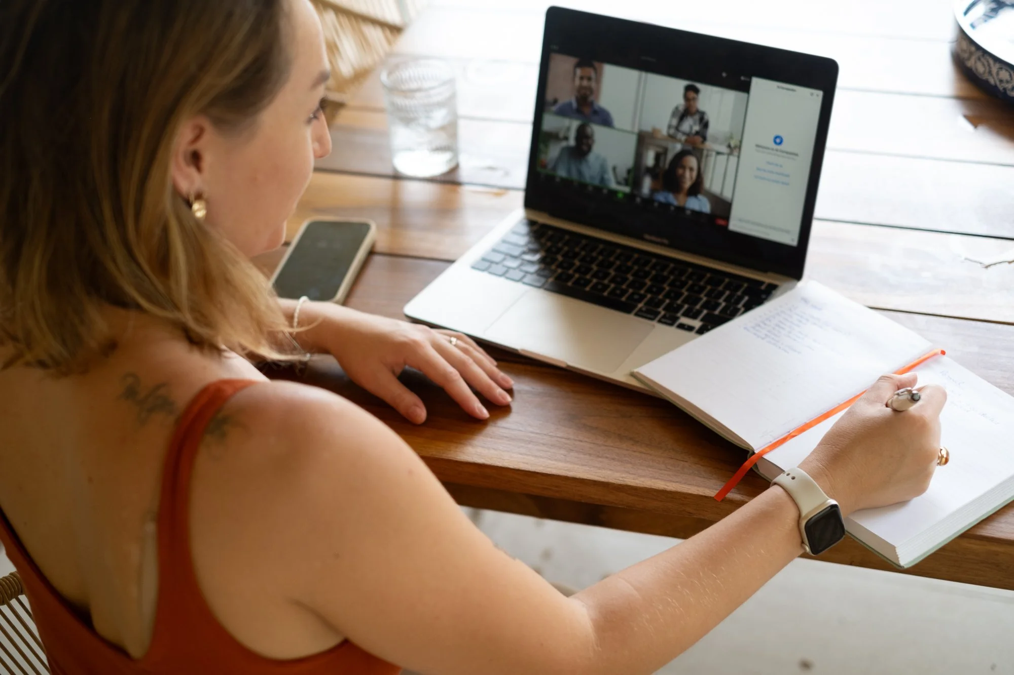 Woman sitting at a wooden table participating in a video conference, taking notes in a notebook, with a smartphone and a glass of water on the table.