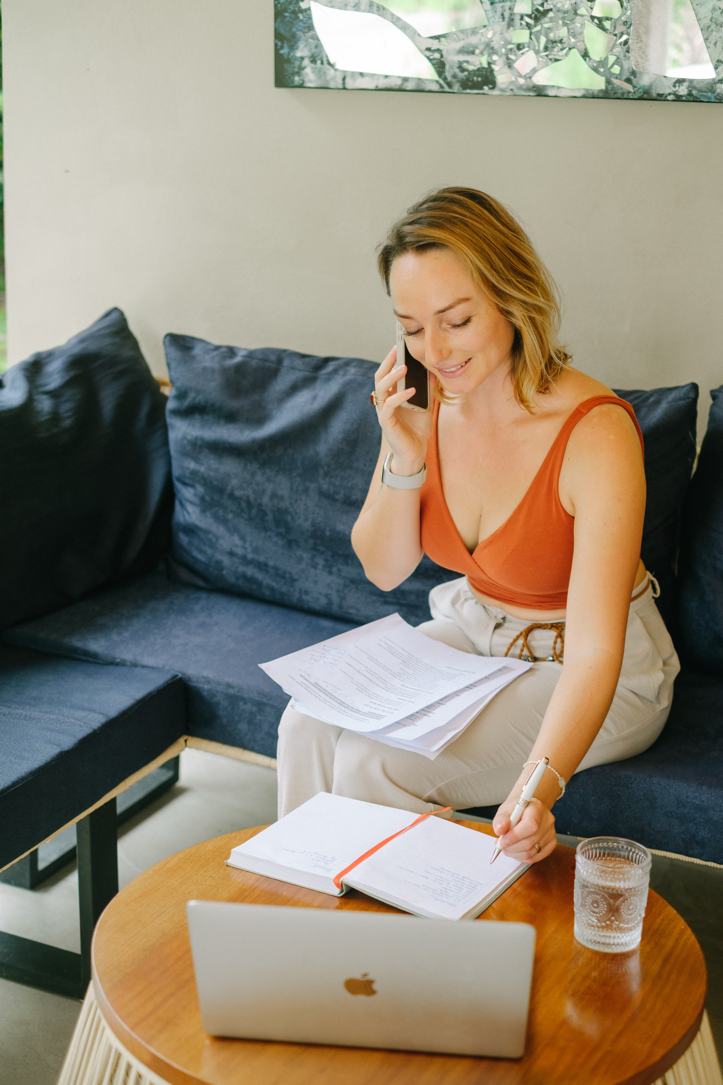 A woman sitting on a dark sofa, talking on her cell phone, with papers, a notebook, and a laptop on a round wooden coffee table in front of her, with a glass of water nearby.