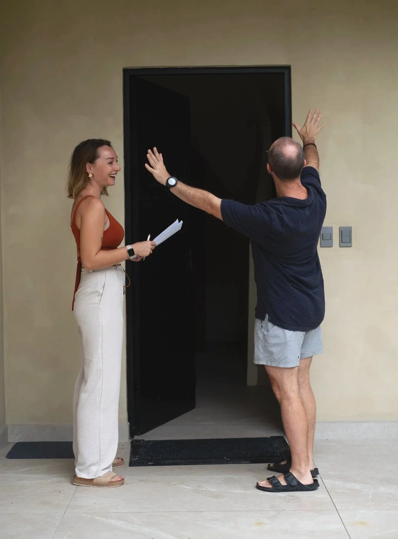 A woman smiling and holding papers, talking to a man in shorts and sandals, standing in front of a dark open door.