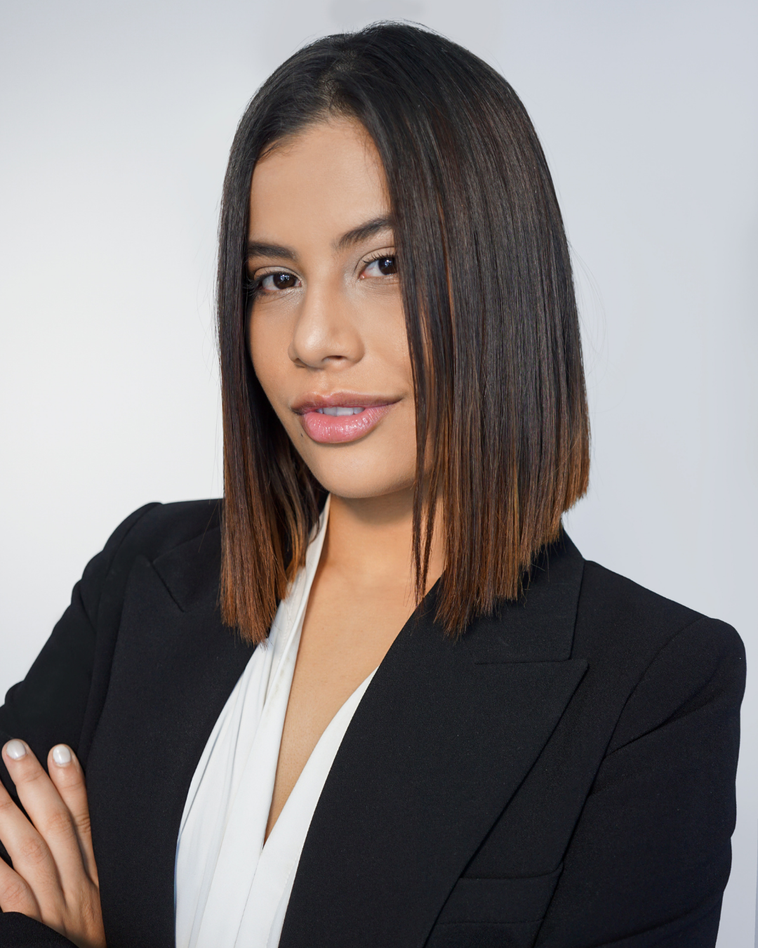 A woman with shoulder-length dark hair with brown highlights, wearing a black blazer and white blouse, posing with arms crossed against a plain light gray background.