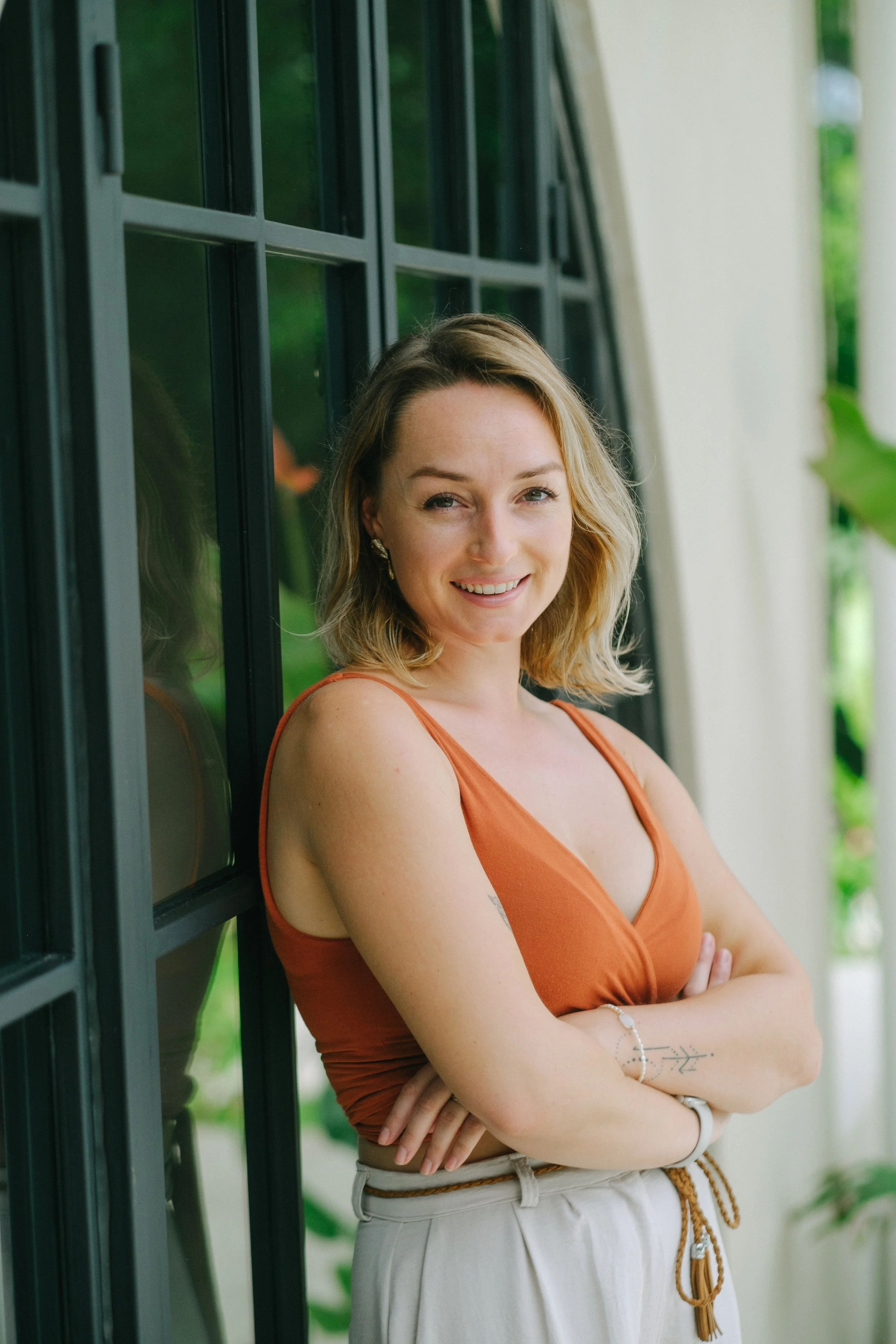 A young woman with shoulder-length blonde hair smiling and crossing her arms, standing outdoors near a black grid-patterned window and greenery.