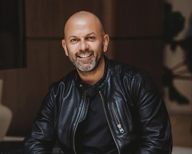 A smiling bald man with a beard wearing a black leather jacket, sitting indoors with a blurred background.