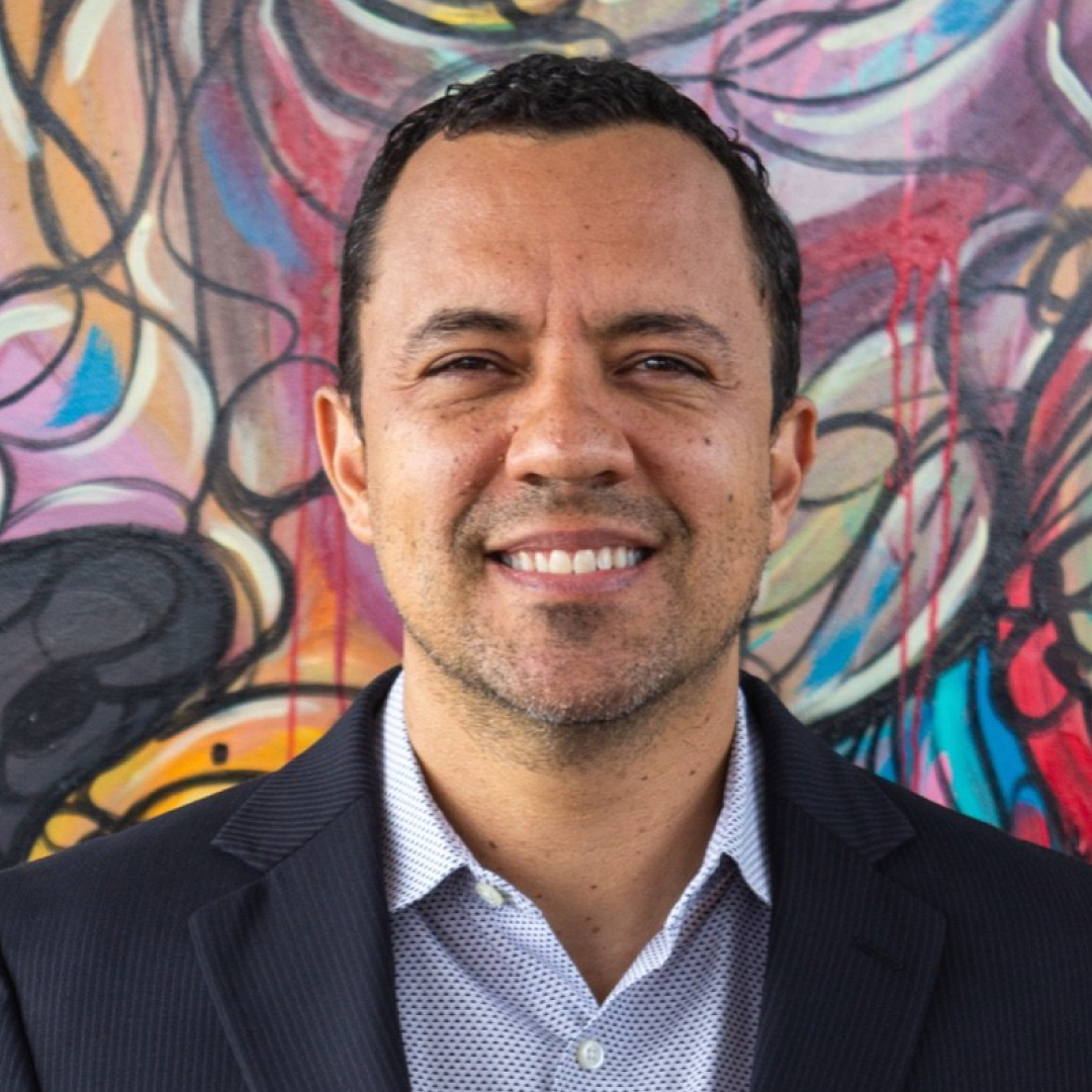 A smiling man in a dark suit and white shirt stands in front of a colorful, abstract mural art background.