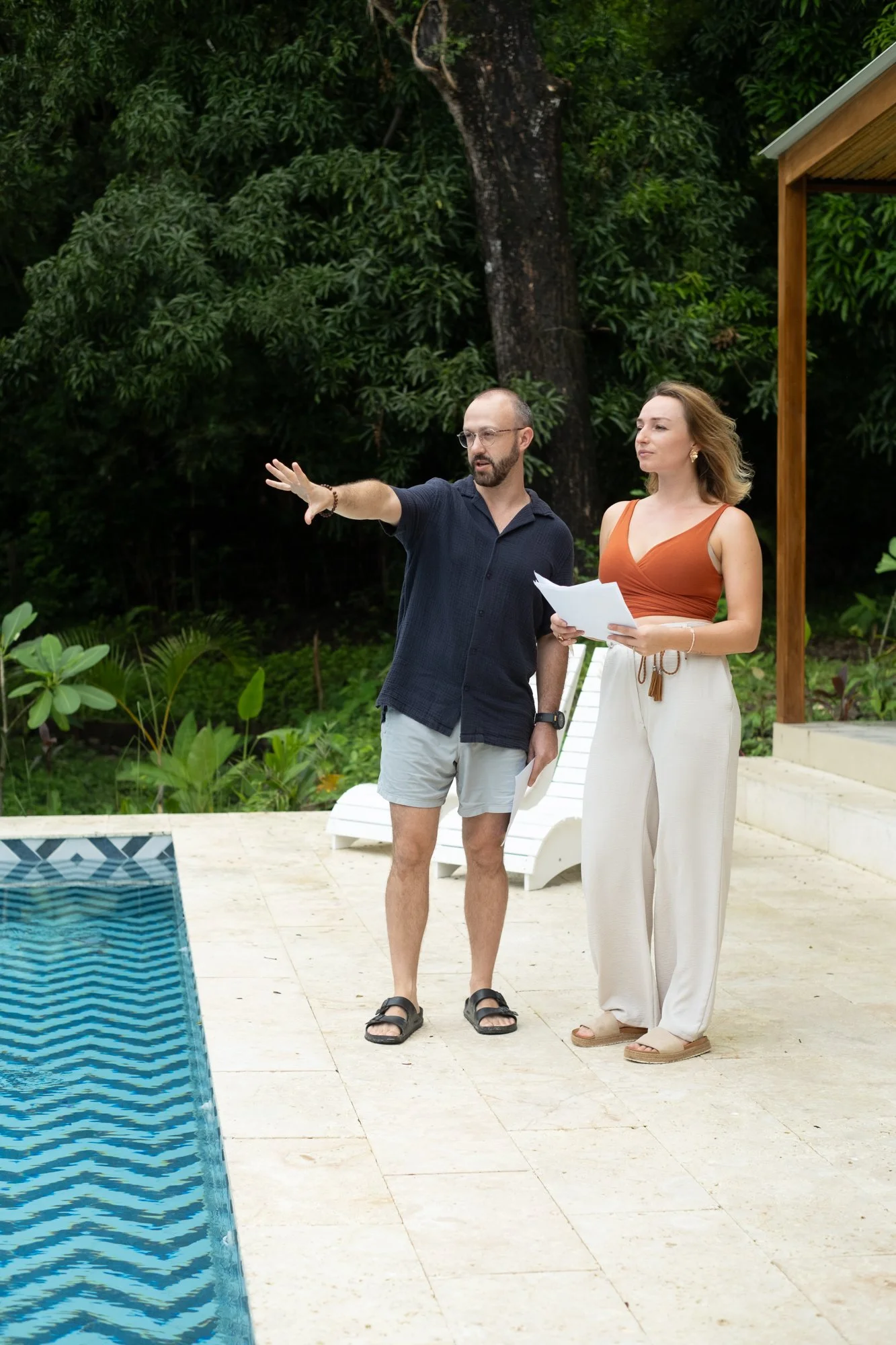 A man and a woman standing by a swimming pool outdoors, looking at each other and discussing. The man is gesturing with his hand, holding papers, while the woman is holding papers. They are dressed casually, near a lush green garden with trees.