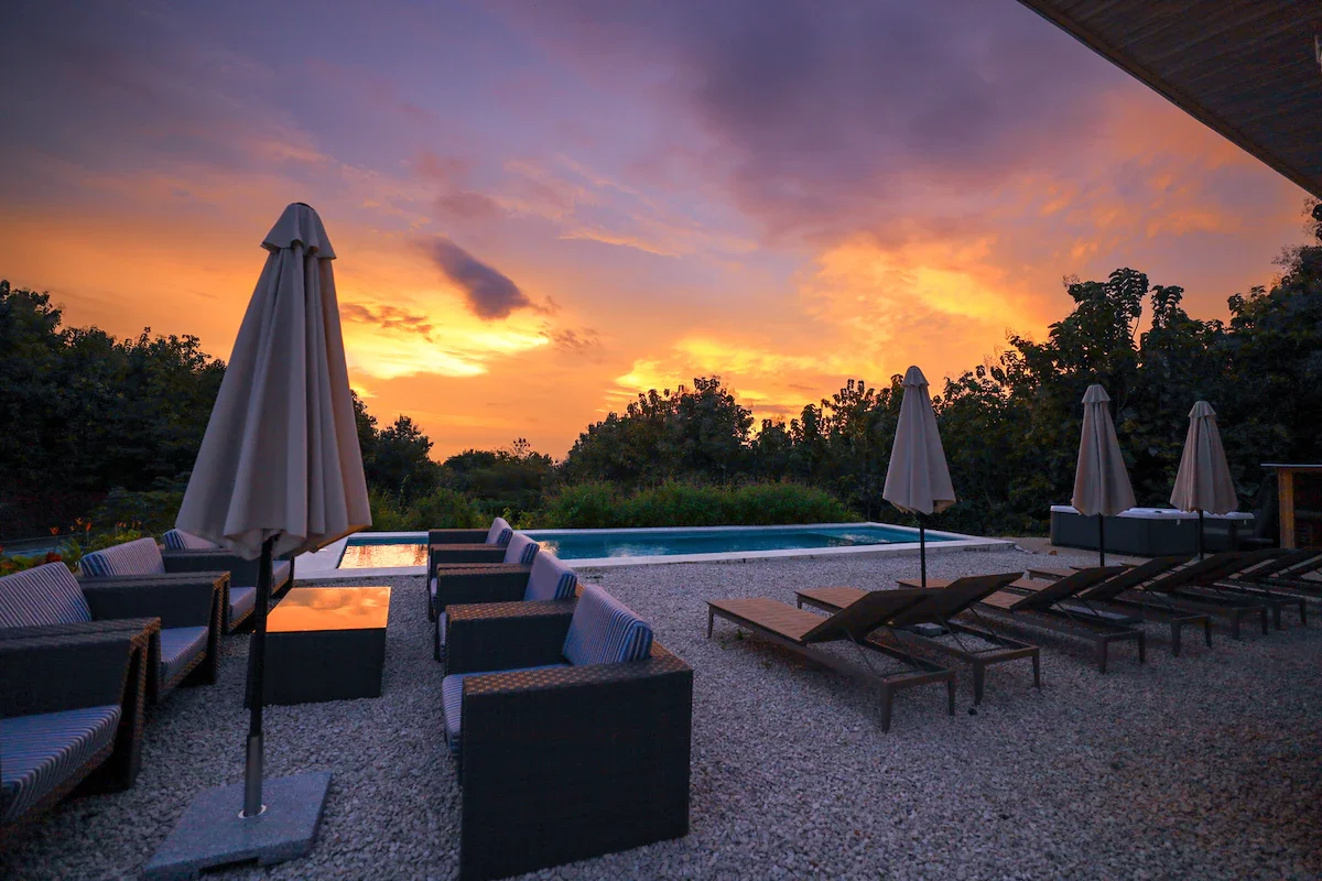 Outdoor poolside lounge area at sunset with umbrellas, chairs, and a view of a colorful sky.