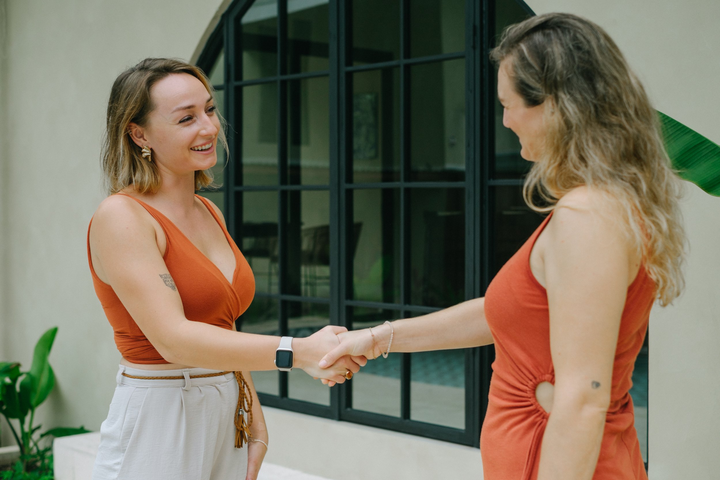 Two women shaking hands and smiling in front of a black window. One woman has shoulder-length hair, wears a sleeveless orange top and cream-colored pants, and the other has long wavy hair, wears an orange top, and has a tattoo on her upper arm.