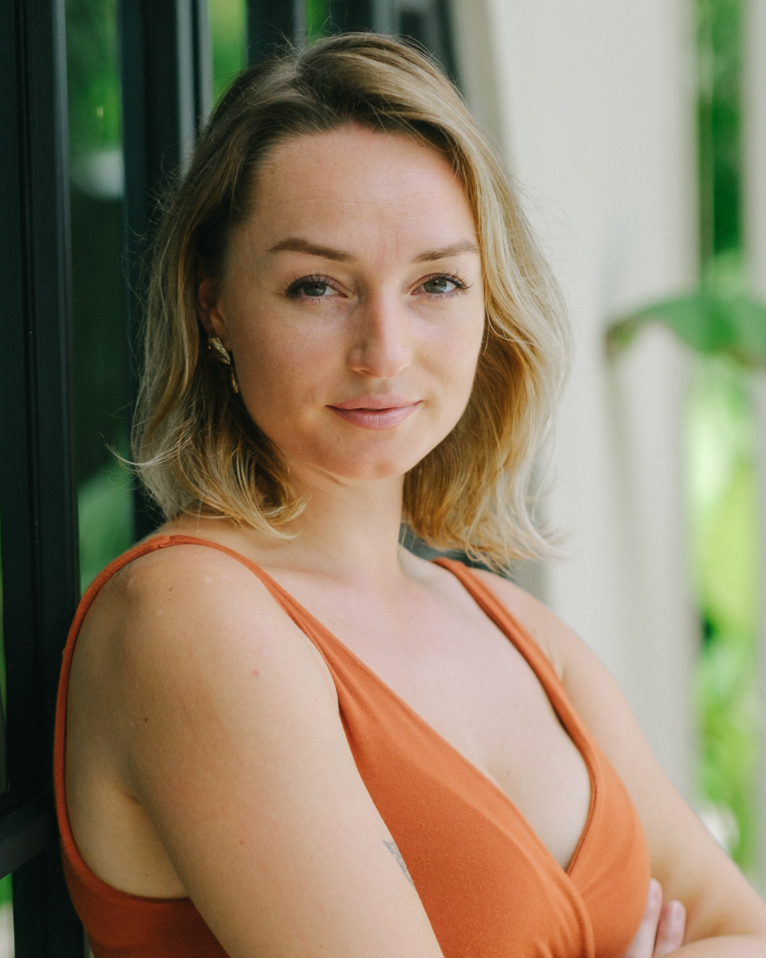 A young woman with shoulder-length wavy blonde hair and light skin, wearing an orange sleeveless top, leaning against a window with a blurred green outdoor background.