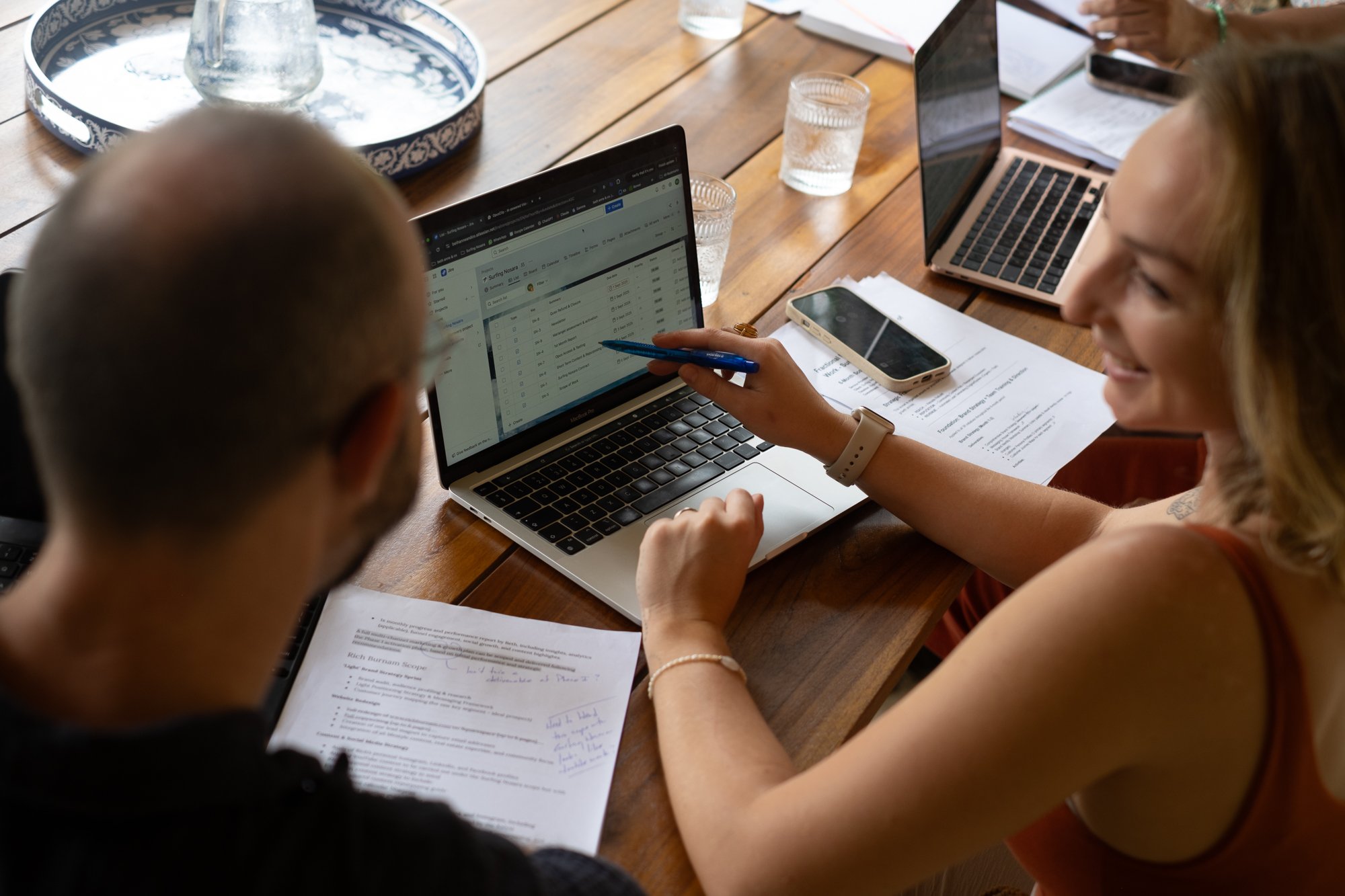 Two women and one man sitting at a wooden table working on laptops and reviewing papers together in a meeting or collaborative workspace.