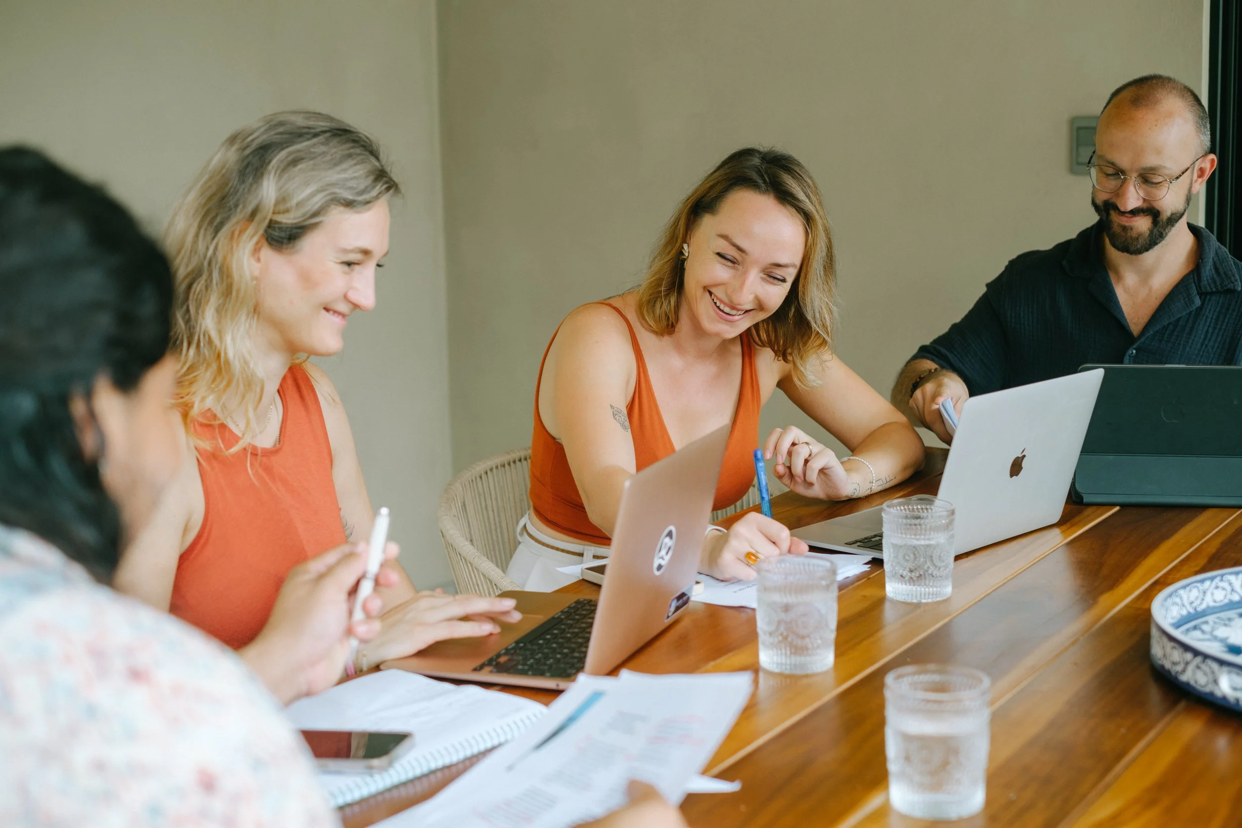 Group of four diverse people sitting around a wooden table, working together with laptops and papers, smiling and enjoying a collaborative meeting.