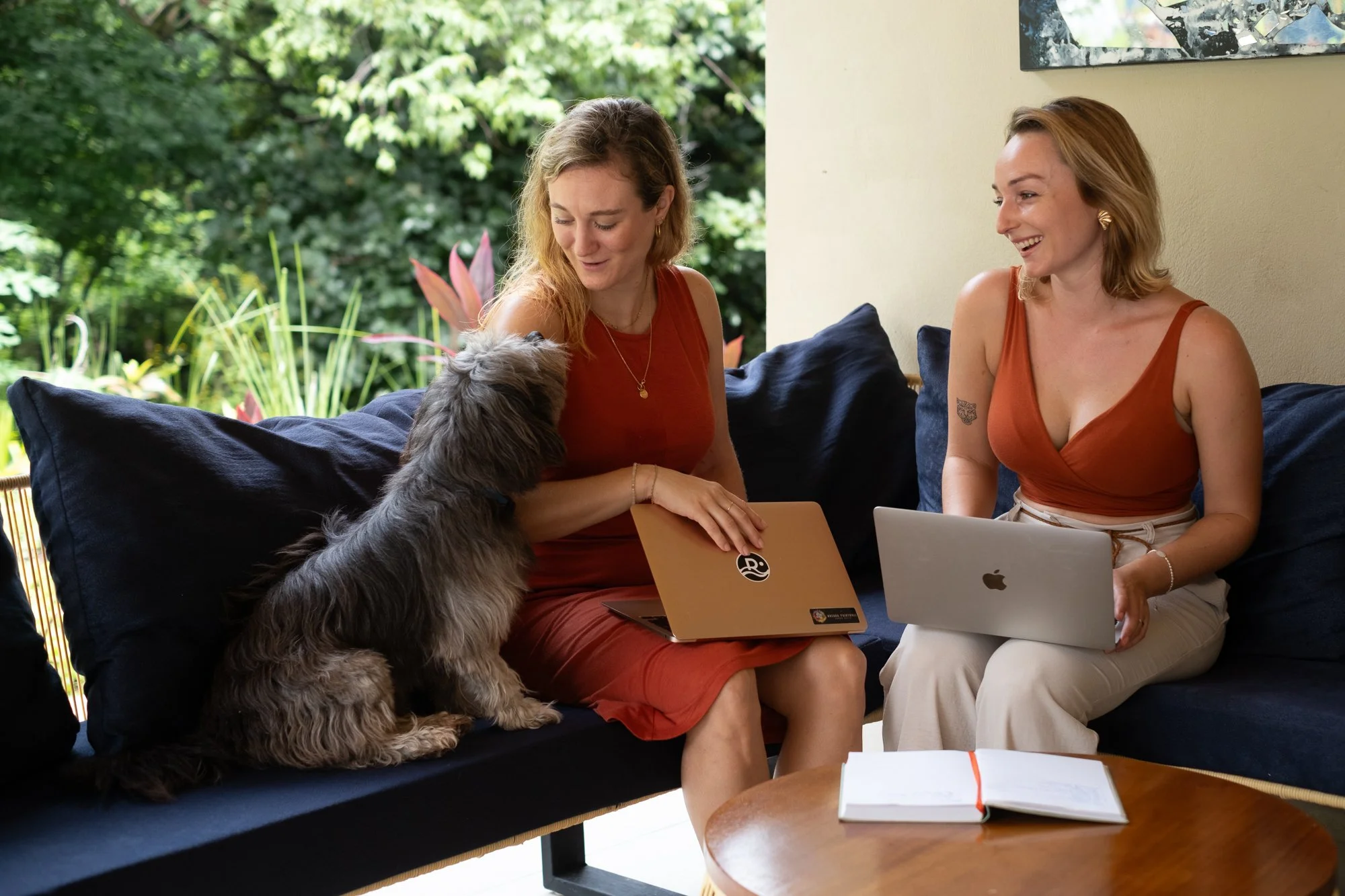 Two women sitting on a dark sofa with a dog between them, working on laptops and smiling, with a green outdoor garden visible through a window behind them.