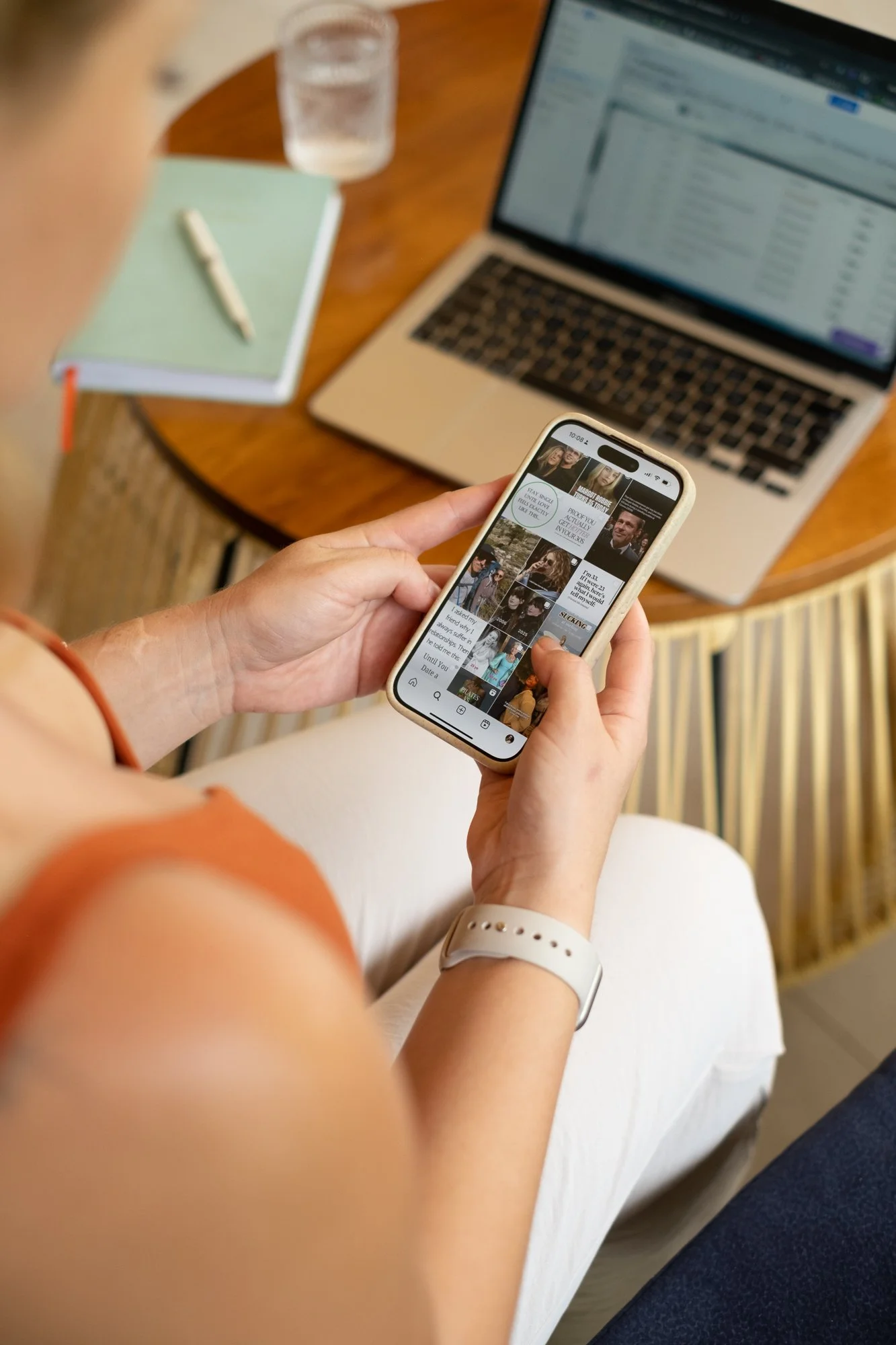 Person holding a smartphone while sitting at a wooden table with a laptop, glass of water, notebook, and pen nearby.