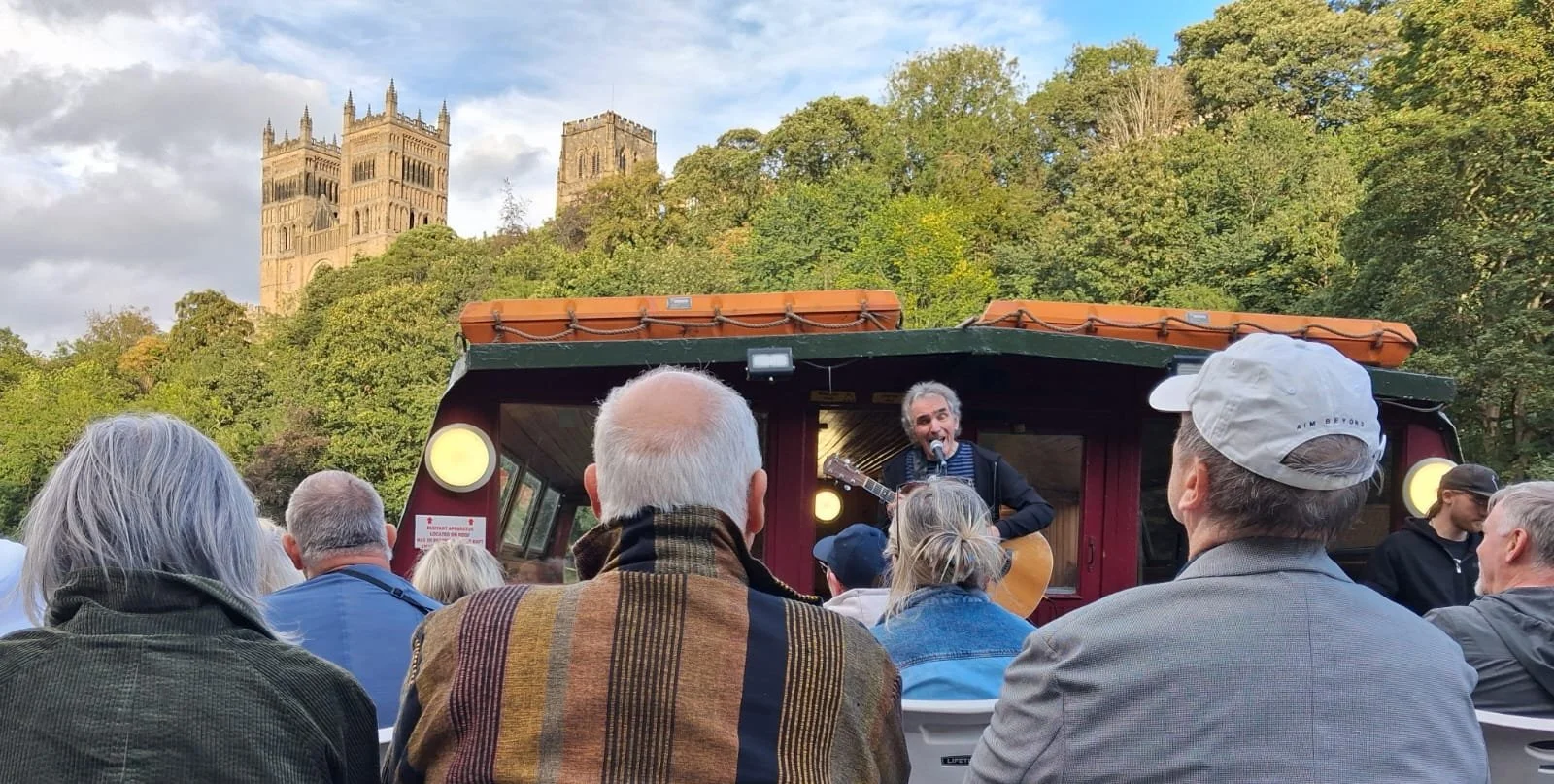 Back view of audience sitting on a boat watching Jez Lowe sing with guitar. Durham Cathedral in background