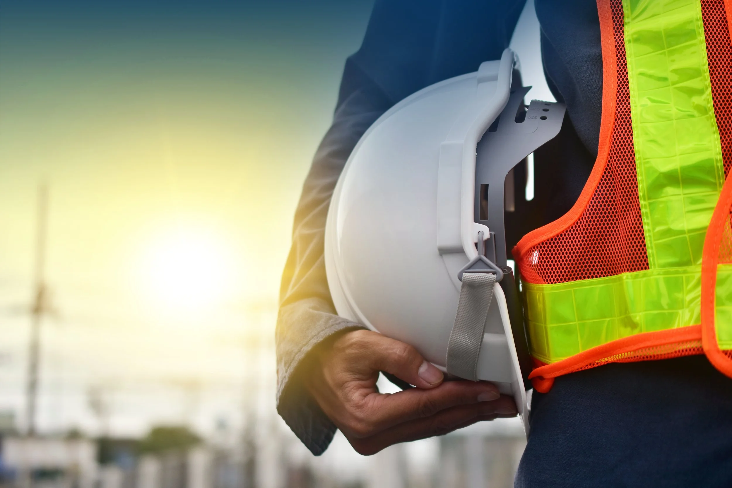 Person in a safety vest holding a white construction helmet, outdoors with a blurred industrial background at sunset.