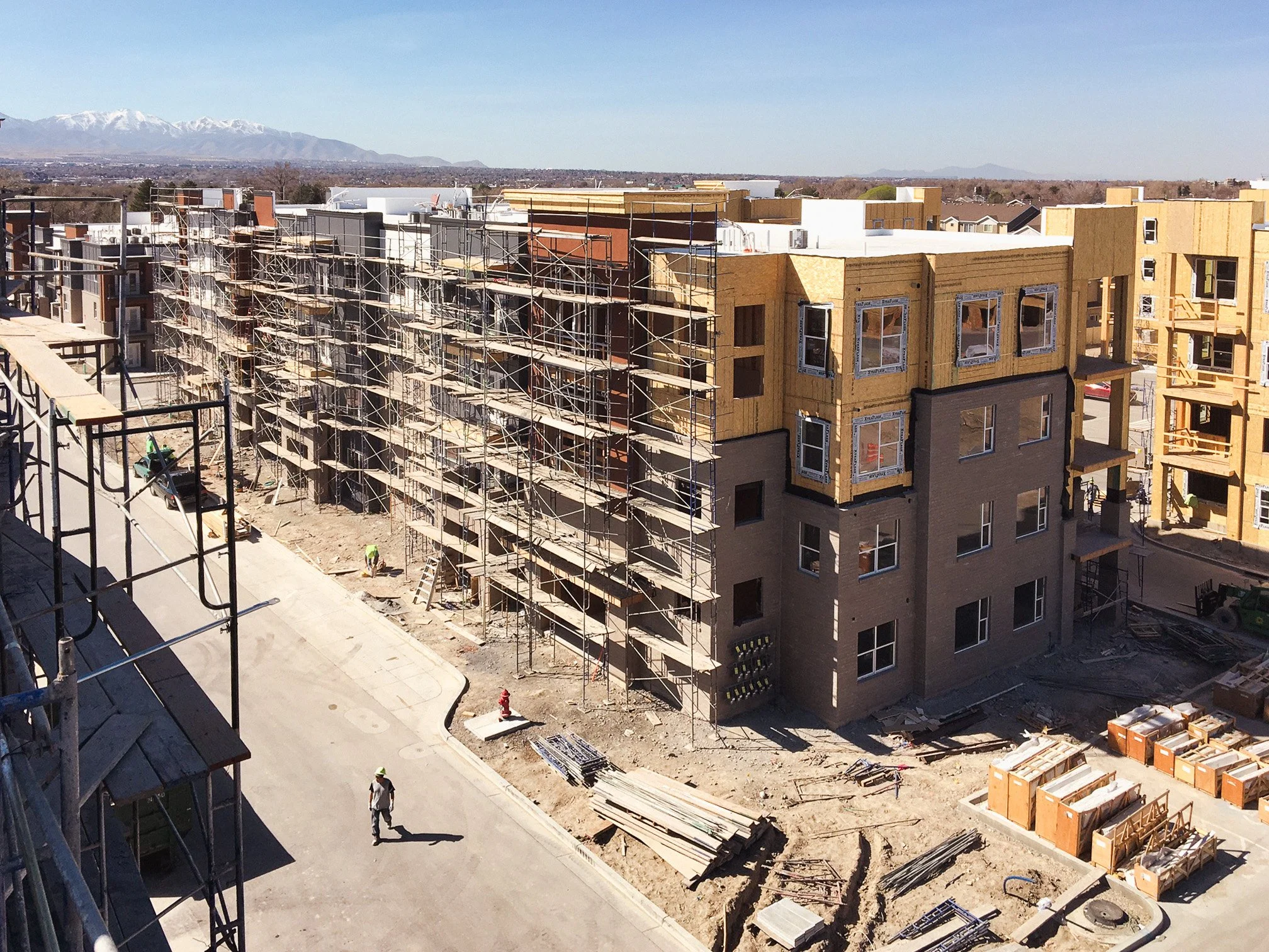 Construction site with partially built apartment buildings, scaffolding, construction workers, and materials, with snow-capped mountains in the background.