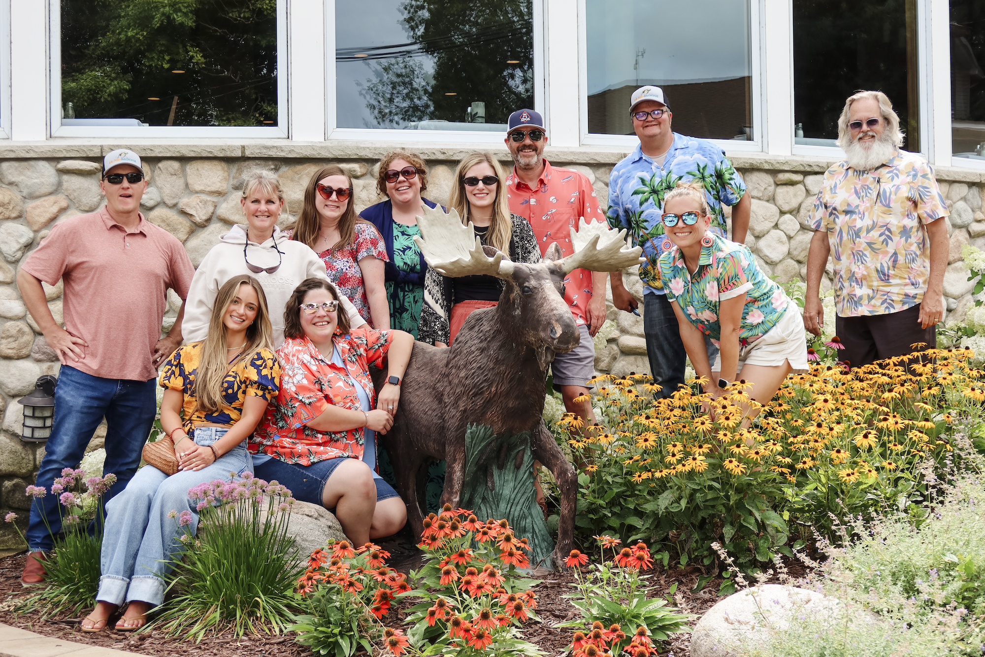 A group of twelve people, including men and women of various ages, poses around a moose statue in a garden with colorful flowers in front of a stone building with large windows.