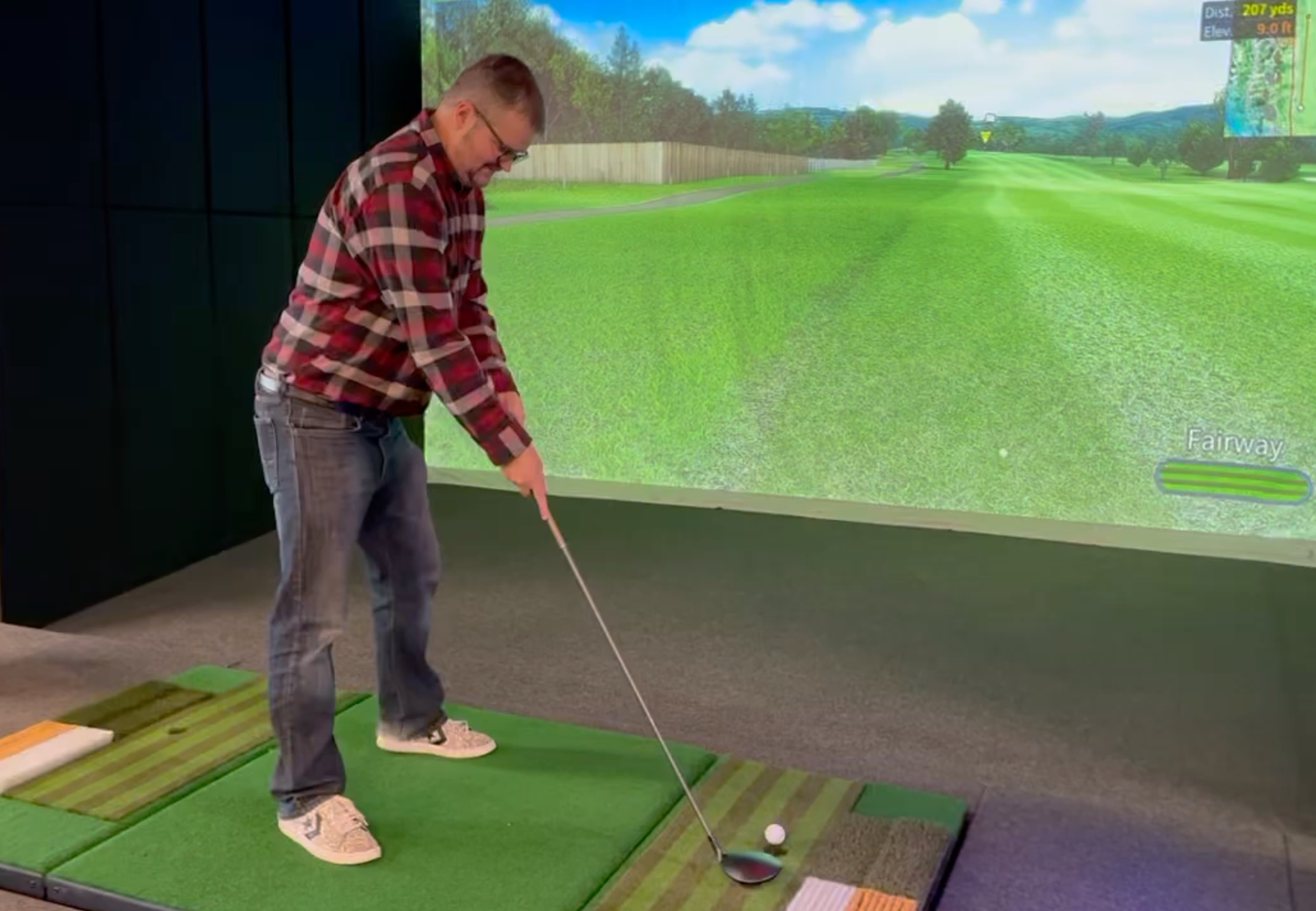 Person practicing golf indoors on a simulated golf course, standing on a mat with a golf club, preparing to hit a golf ball.