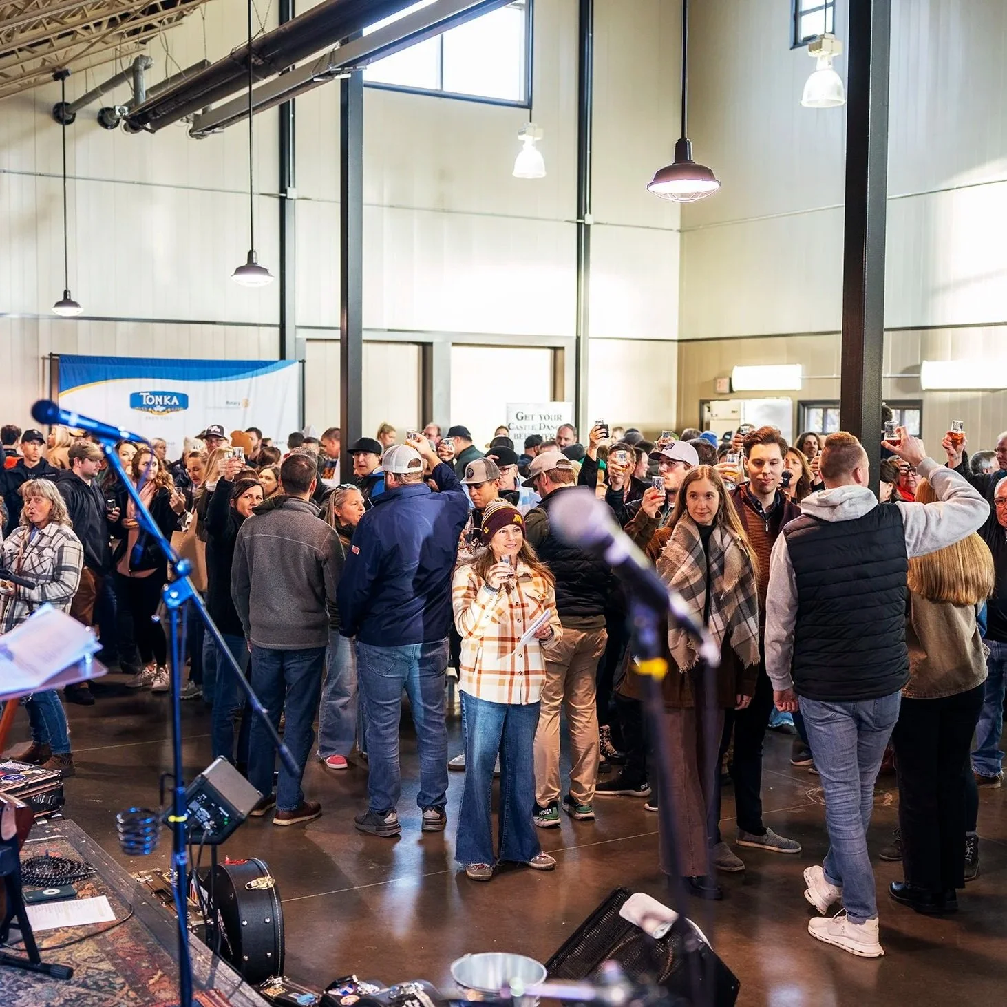 People gathered in an indoor event space, socializing and holding drinks, with a microphone and DJ equipment in the foreground.