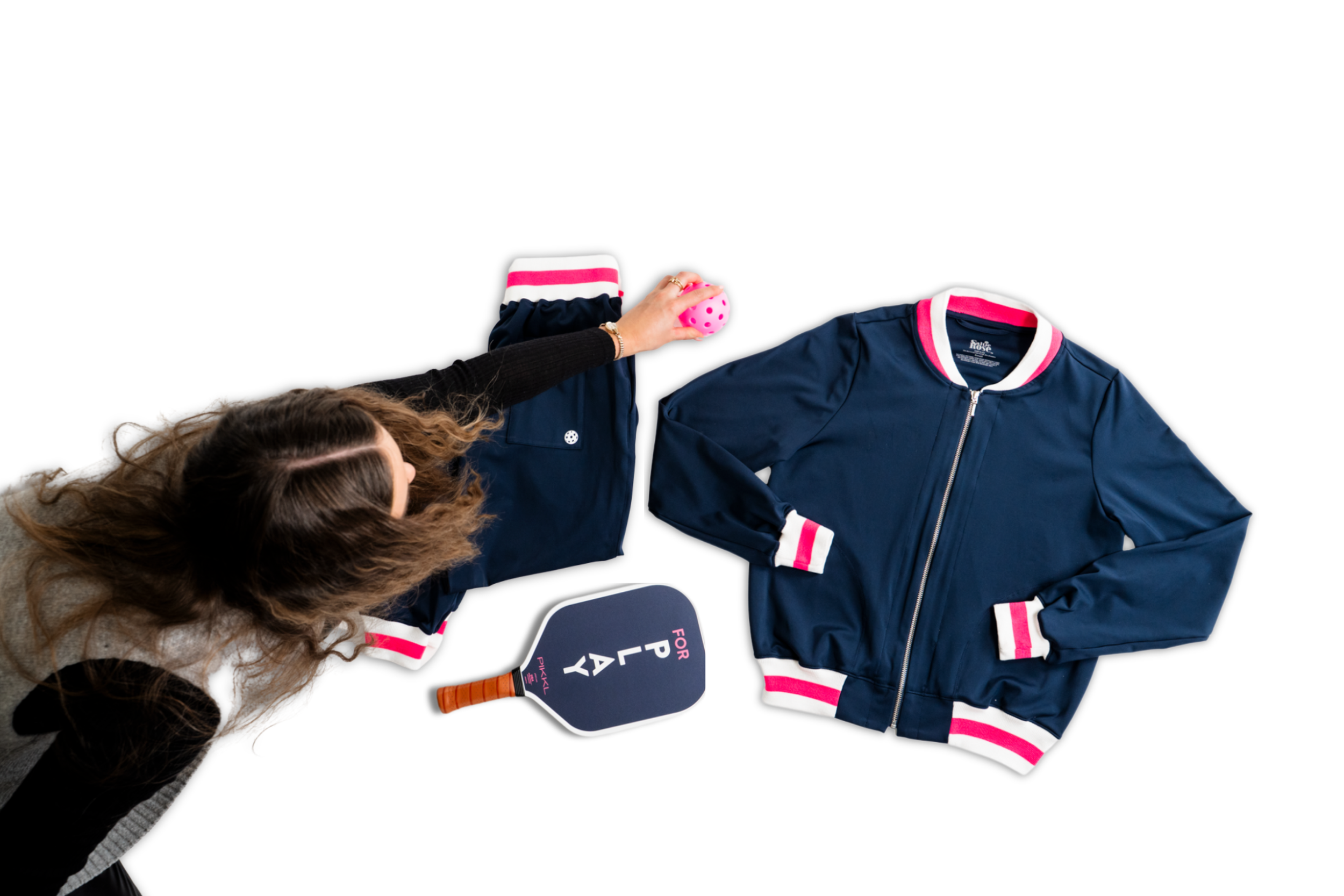 A woman with long, curly hair reaching for a pink tennis ball. Items on a white background include a navy blue and pink athletic jacket, navy blue shorts, and a ping pong paddle with words "FOR PLAY" on the surface.