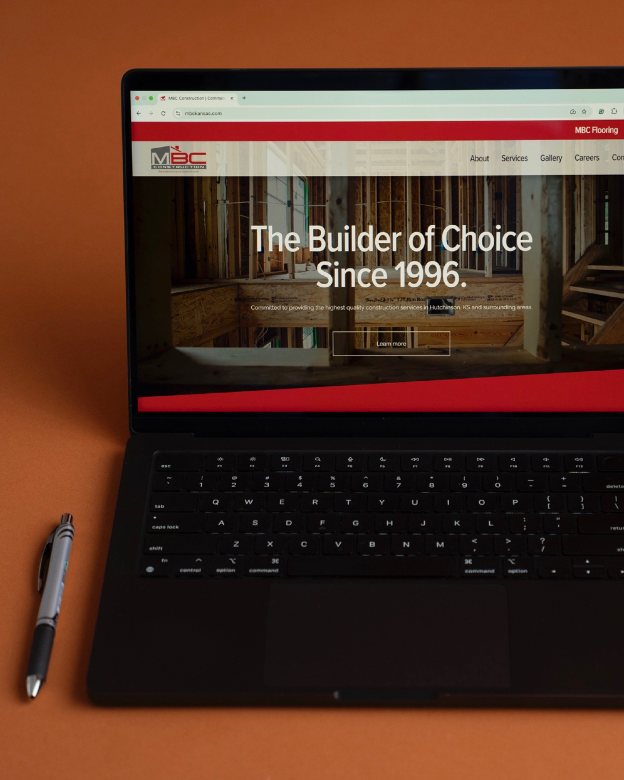 A laptop on an orange surface displaying a construction company's website with the text 'The Builder of Choice Since 1996' and a pen next to it.